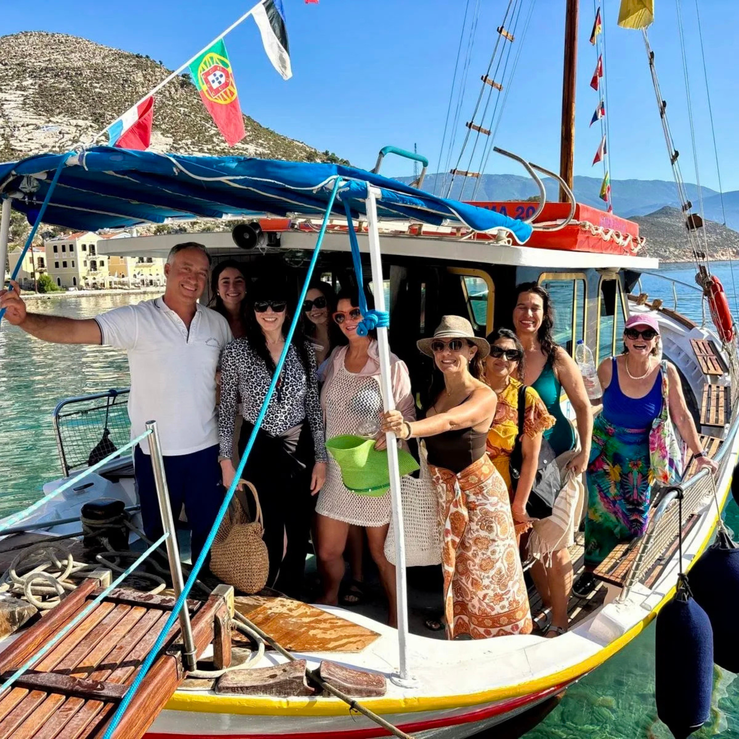 A group of people on a boat in a harbor with mountains in the background, smiling and wearing summer clothing.