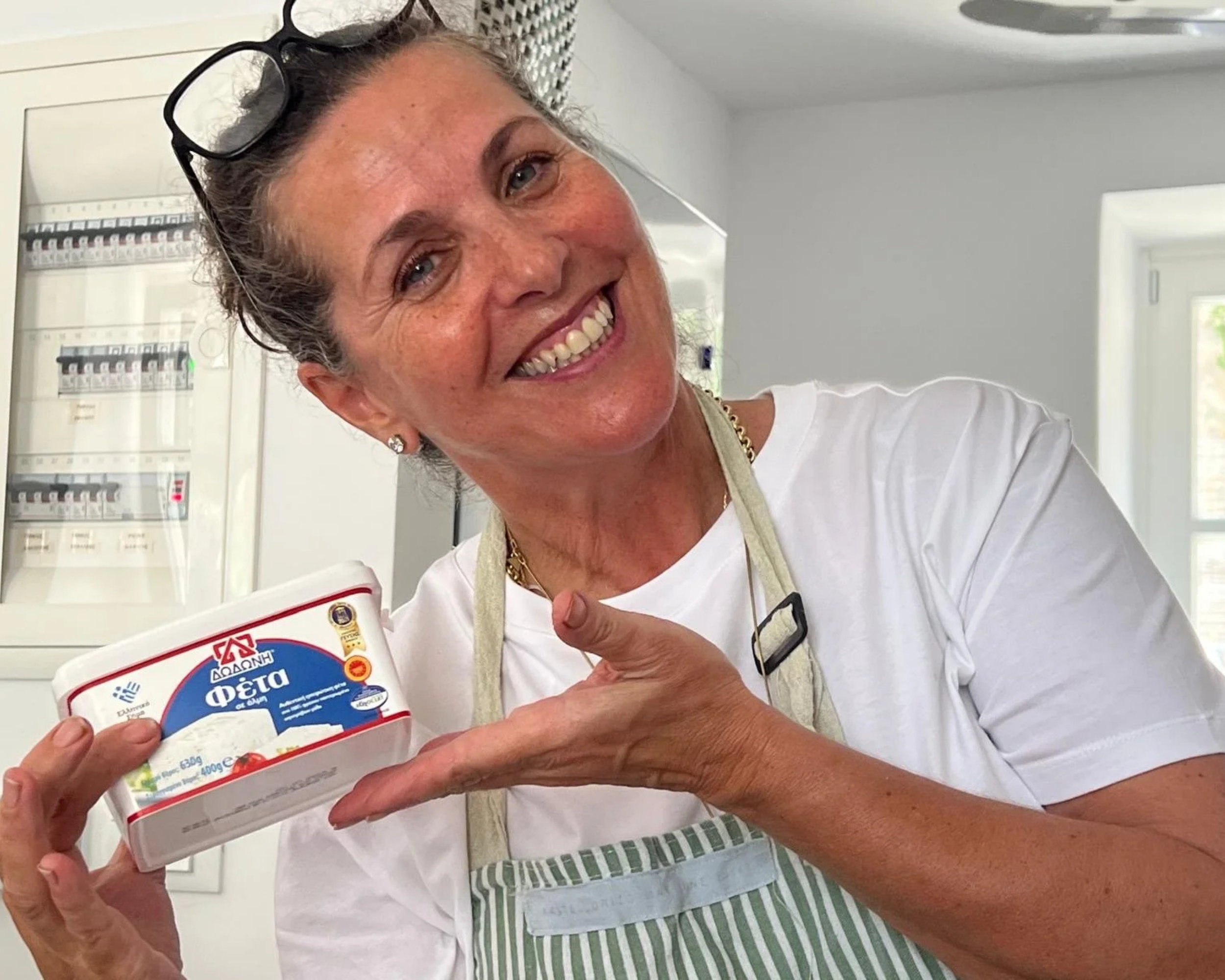 A smiling woman in a white shirt and striped apron holding a container of Greek feta cheese, pointing at it, in a kitchen setting.