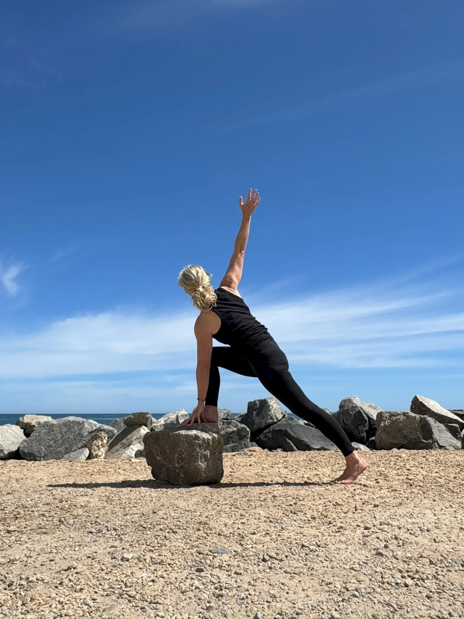 A woman practicing yoga outdoors on a rocky beach during the day. She is in a side plank pose with her left hand on the ground and her right arm extended upward. The sky is clear with some clouds.