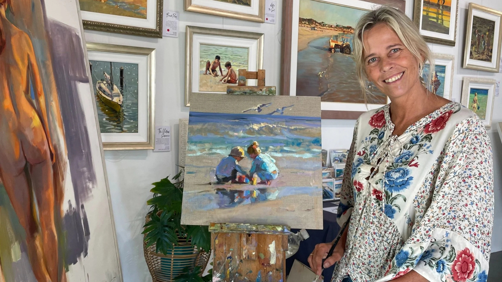 A woman smiling at an art gallery, standing next to a painting of two children sitting on the beach, with various framed landscape and seascape paintings hanging on the wall behind her.