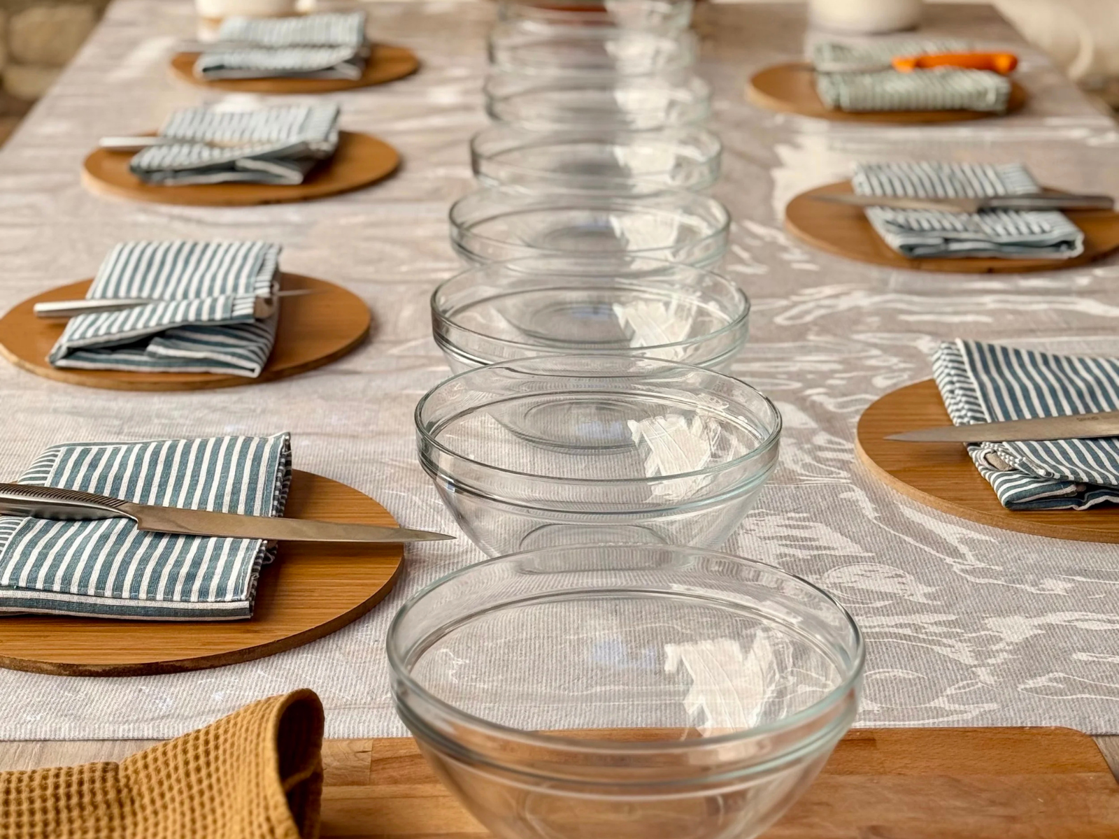 A dining table set with empty glass bowls, striped cloth napkins on wooden chargers, and cutlery, arranged on a white tablecloth.