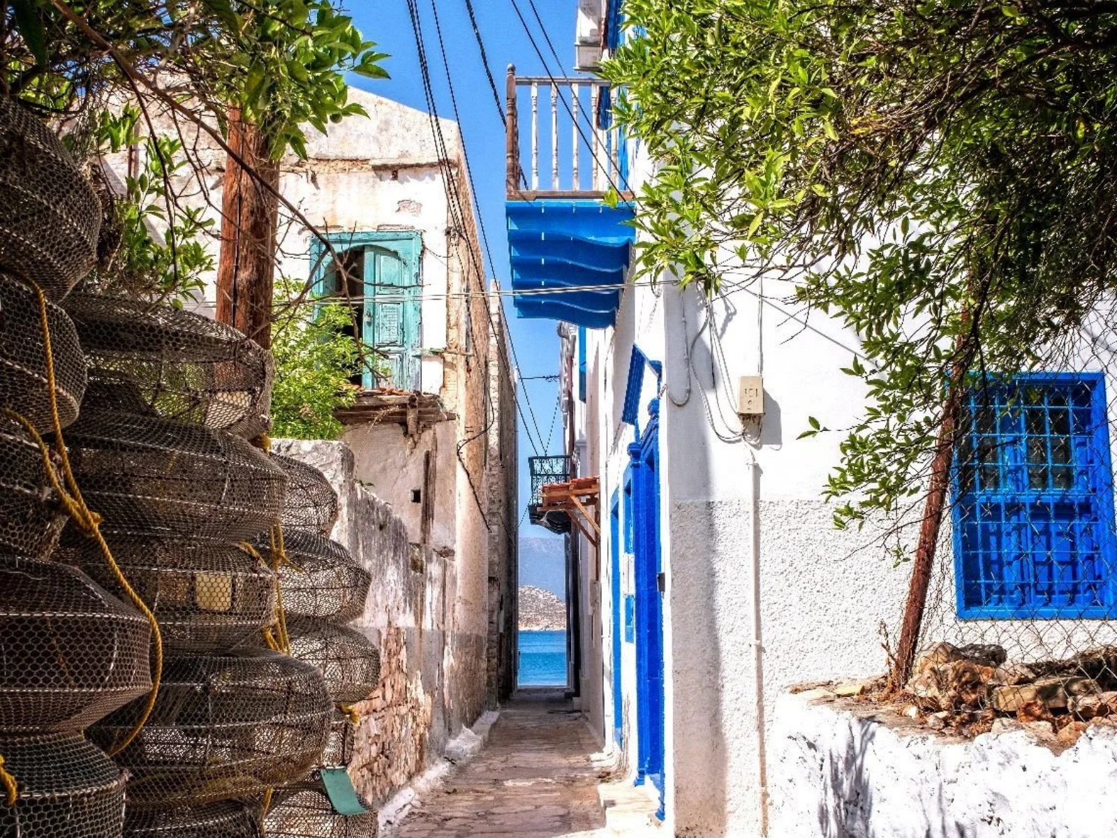 Narrow alleyway in a Mediterranean village with white walls and blue window frames, featuring stacked lobster traps on the left and leafy trees casting shadows on the right.