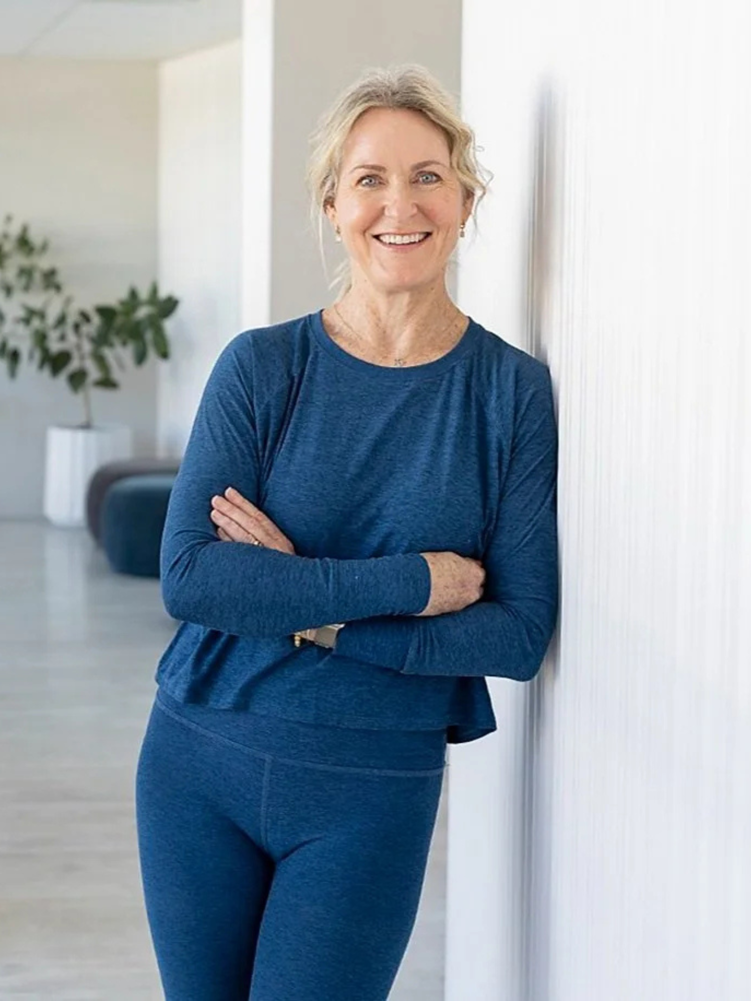 A smiling woman with blonde hair in a casual blue outfit standing indoors against a white wall, with a houseplant in a white pot in the background.