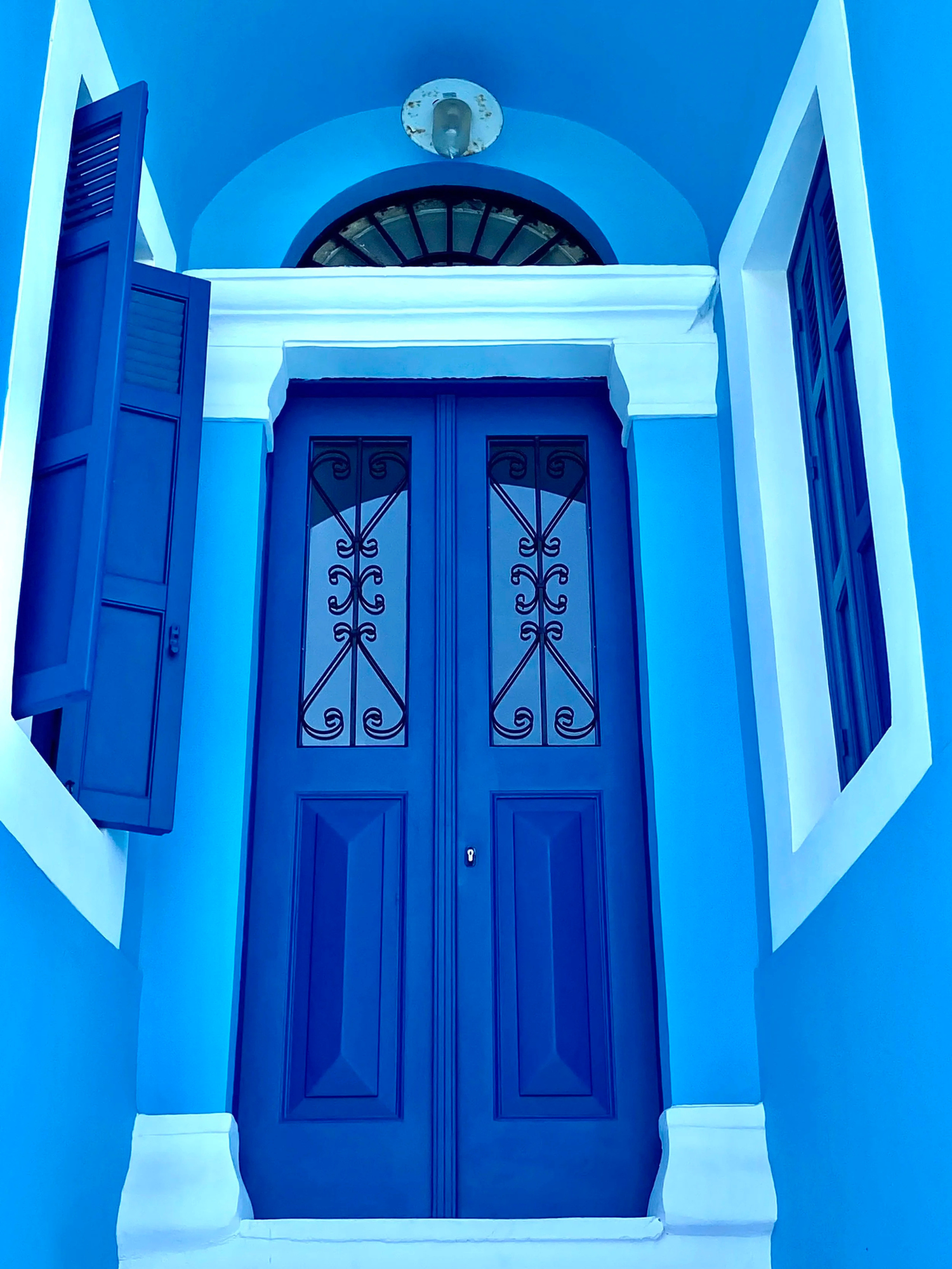 Blue double door with black wrought iron grillwork, white trim, flanked by windows with blue shutters, blue walls, arched window above, and white baseboards.