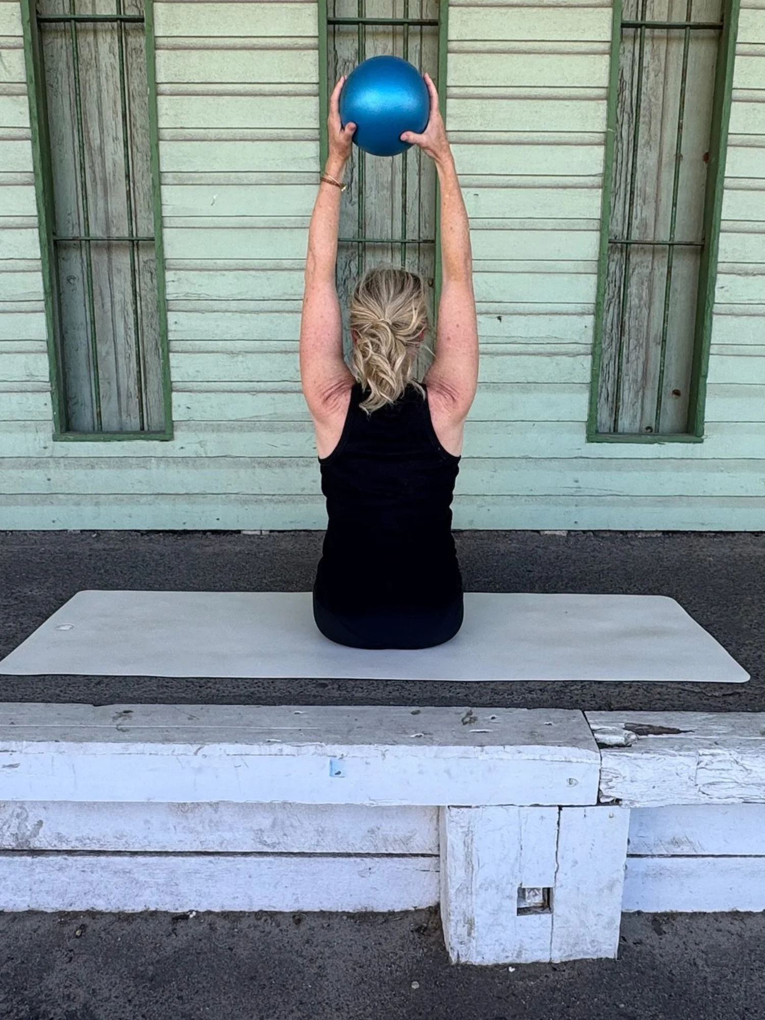 A person with blonde hair tied back, wearing a black sleeveless top, is sitting on a white wooden bench outdoors, holding a blue exercise ball overhead with both hands.