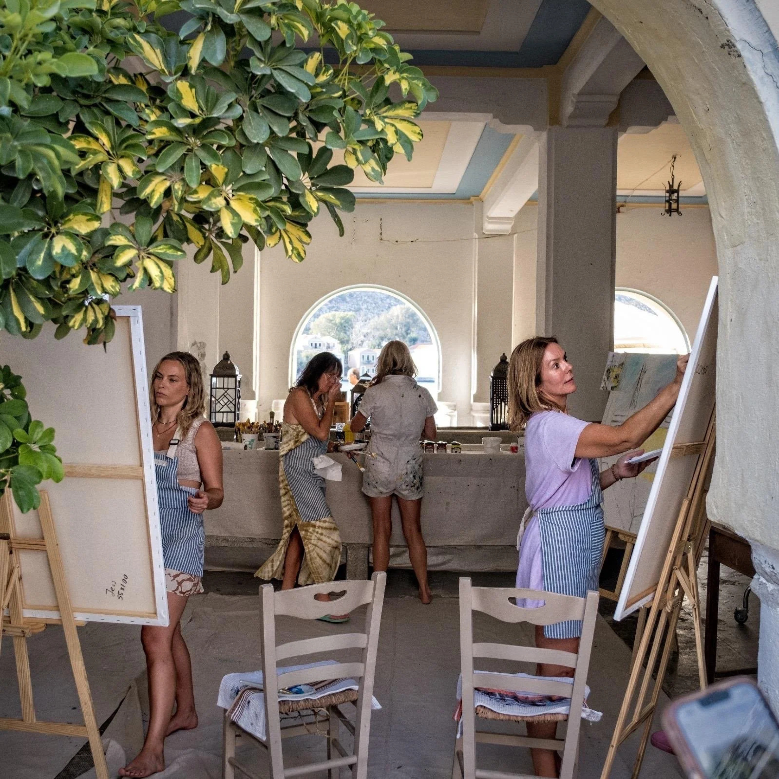 Women painting on canvases in an art class set in a bright room with large arched windows and a white wall, some women are working at a countertop with art supplies, and a large leafy plant is hanging from the ceiling.