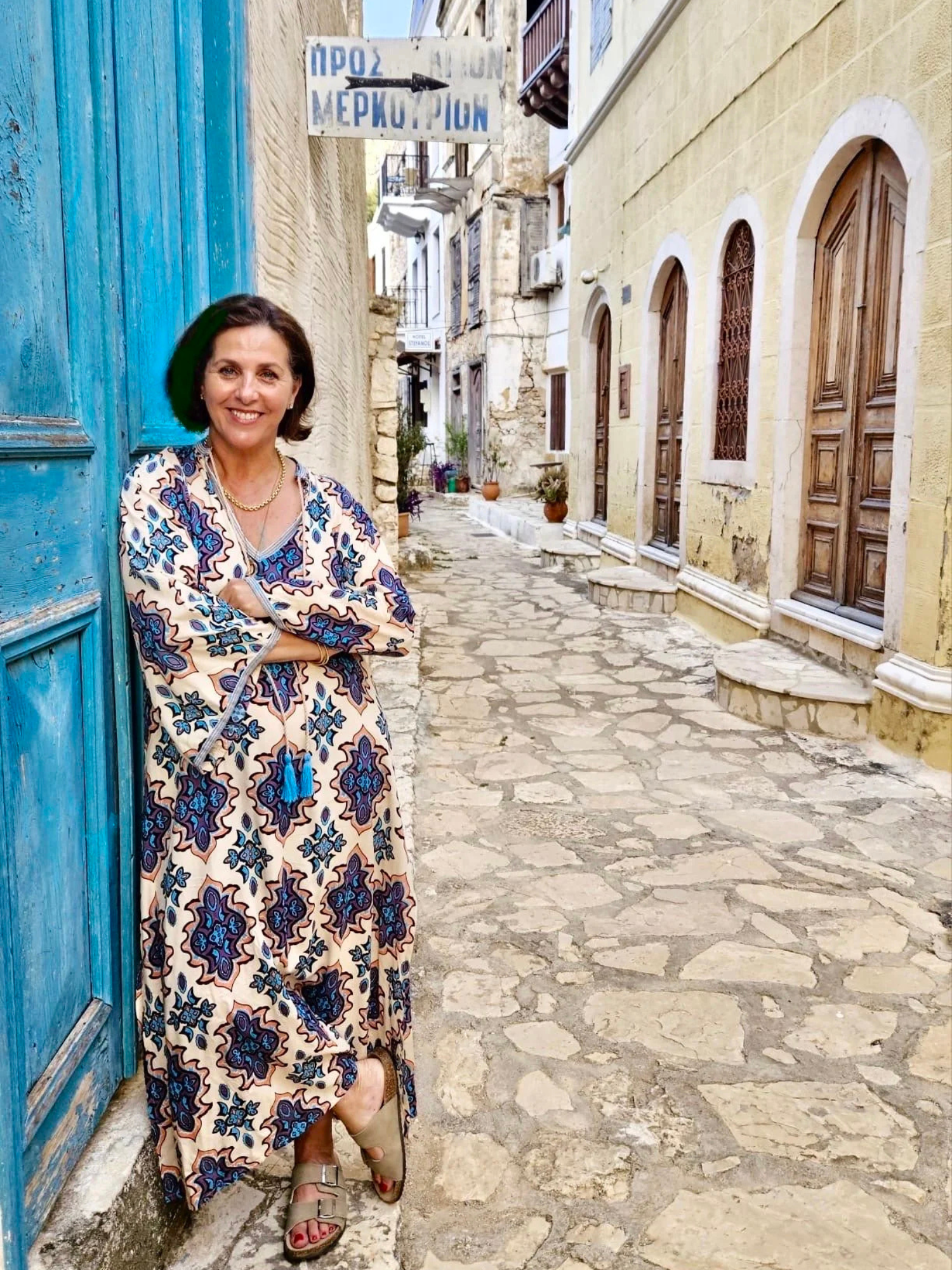 A woman in a patterned dress standing on a cobblestone street next to a blue door, with old buildings and potted plants in the background.