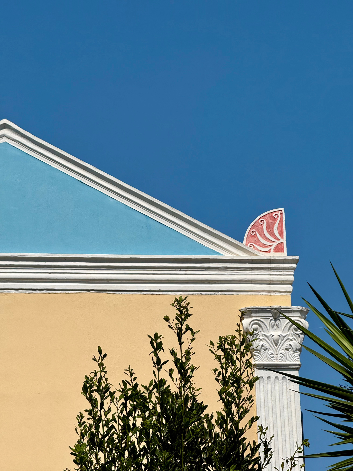 A colorful building with pastel blue, yellow, and pink accents, and architectural column details, set against a clear blue sky with some greenery in the foreground.
