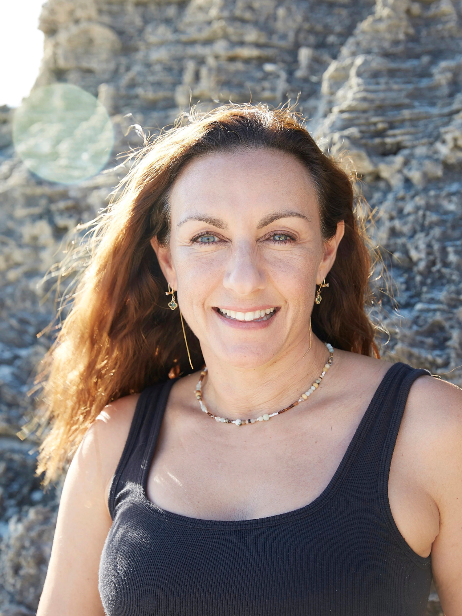 A woman with wavy brown hair and blue eyes smiling outdoors against a rocky background, wearing a black tank top, earrings, and a necklace.
