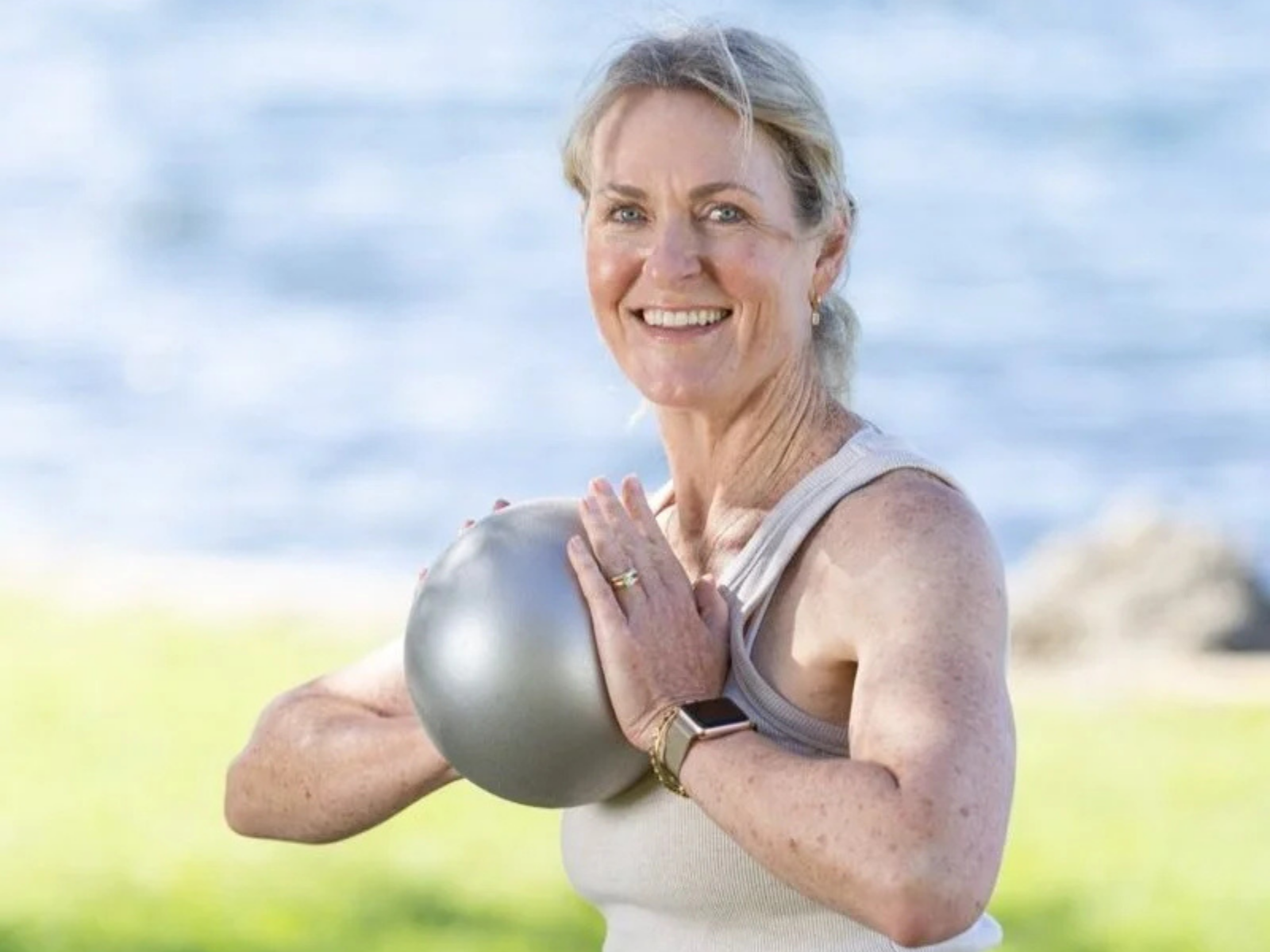 Older woman with gray hair smiling while holding a kettlebell outdoors near water