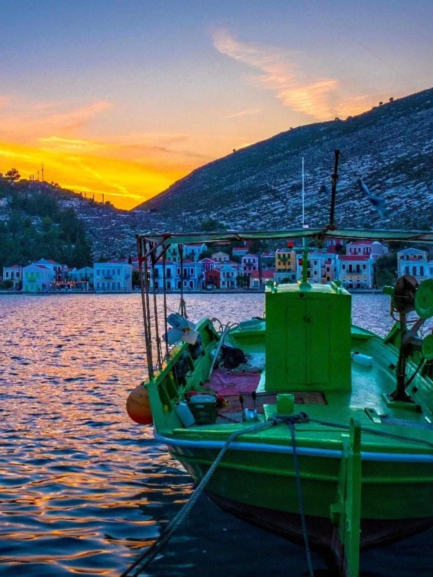 A green boat docked on a calm lake at sunset with colorful buildings on the opposite shore and a mountain in the background.