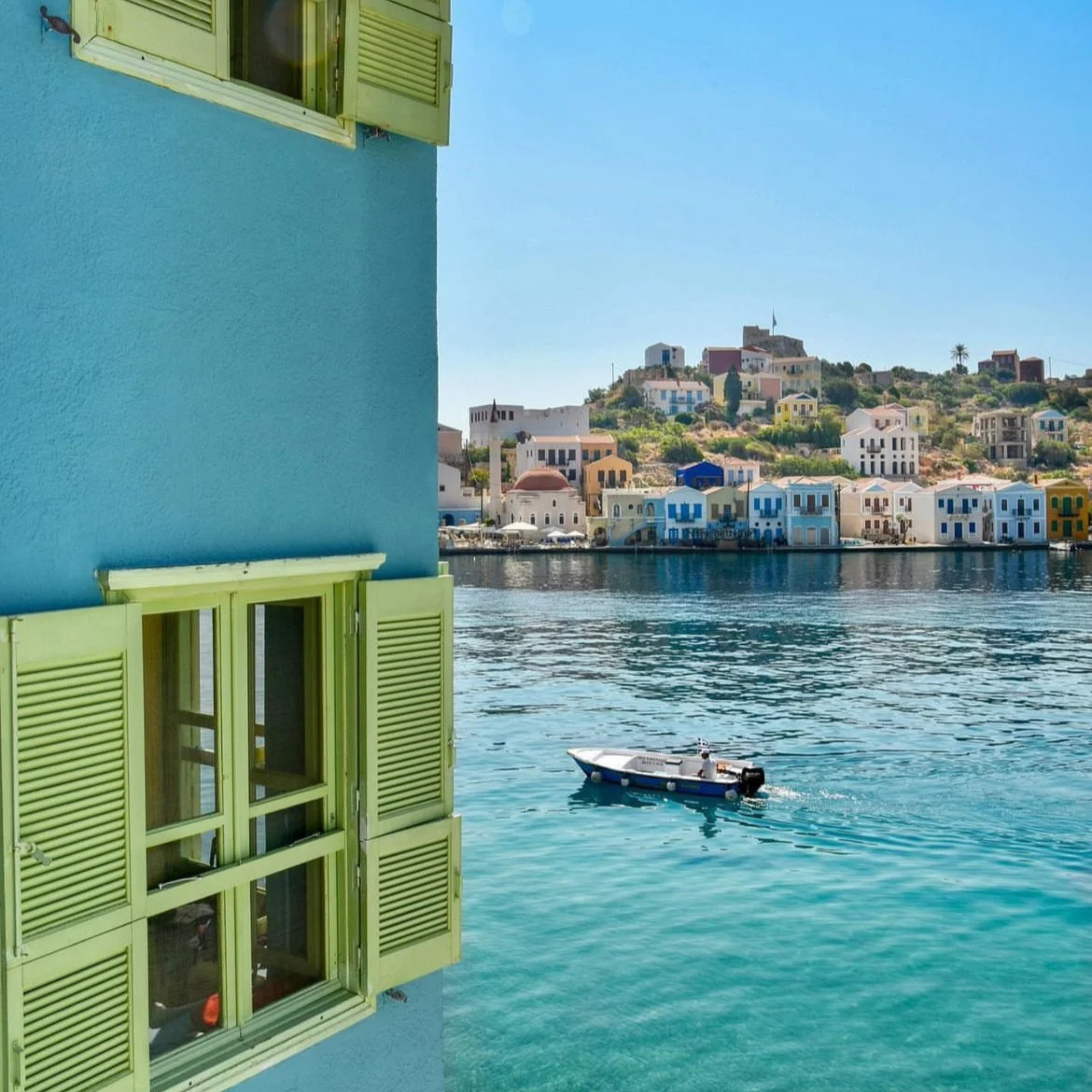 Blue building with lime green shutters near turquoise water, with a small boat and colorful buildings on a hill in the background.