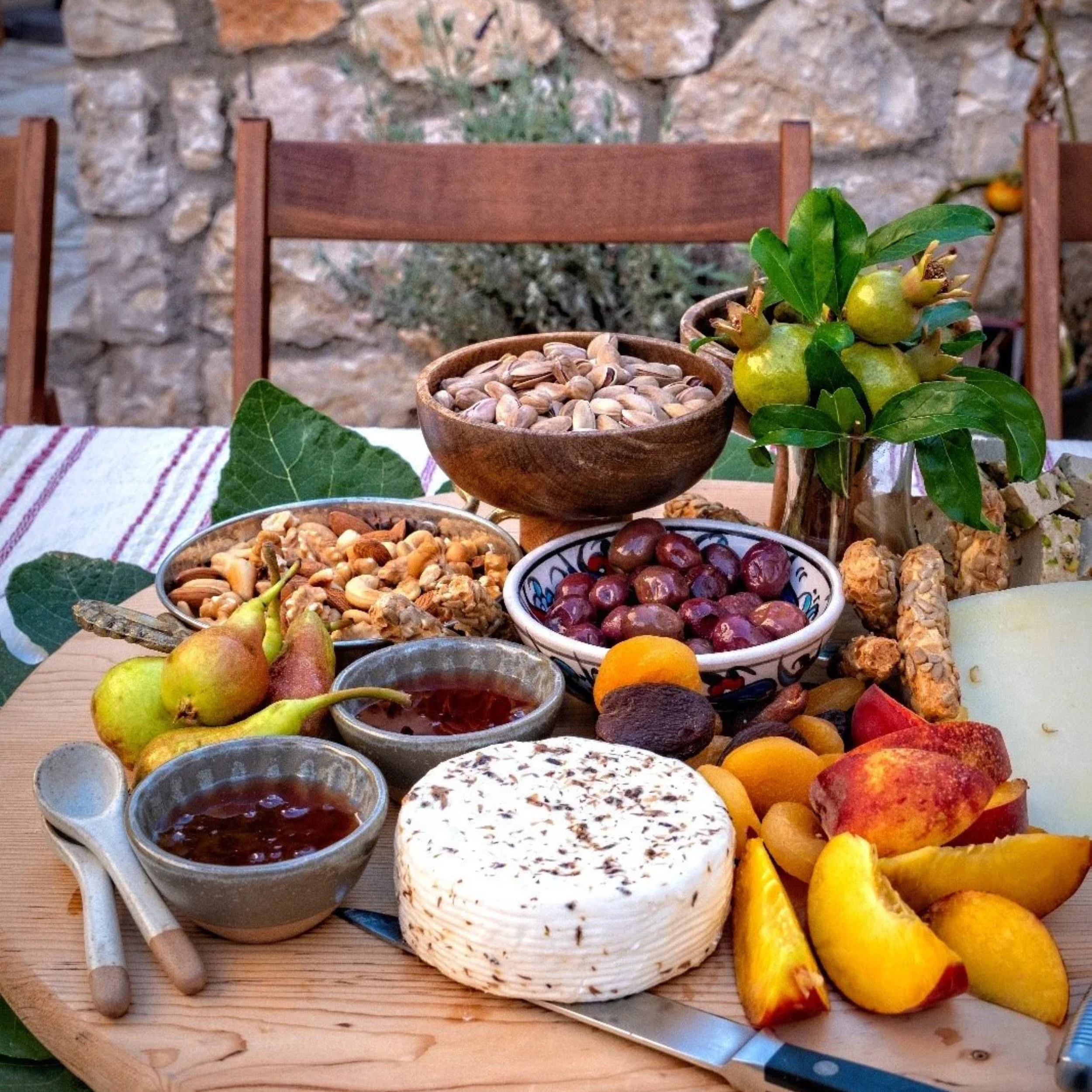 A rustic table with cheese, fresh fruit, nuts, and jams, set against a stone wall background.
