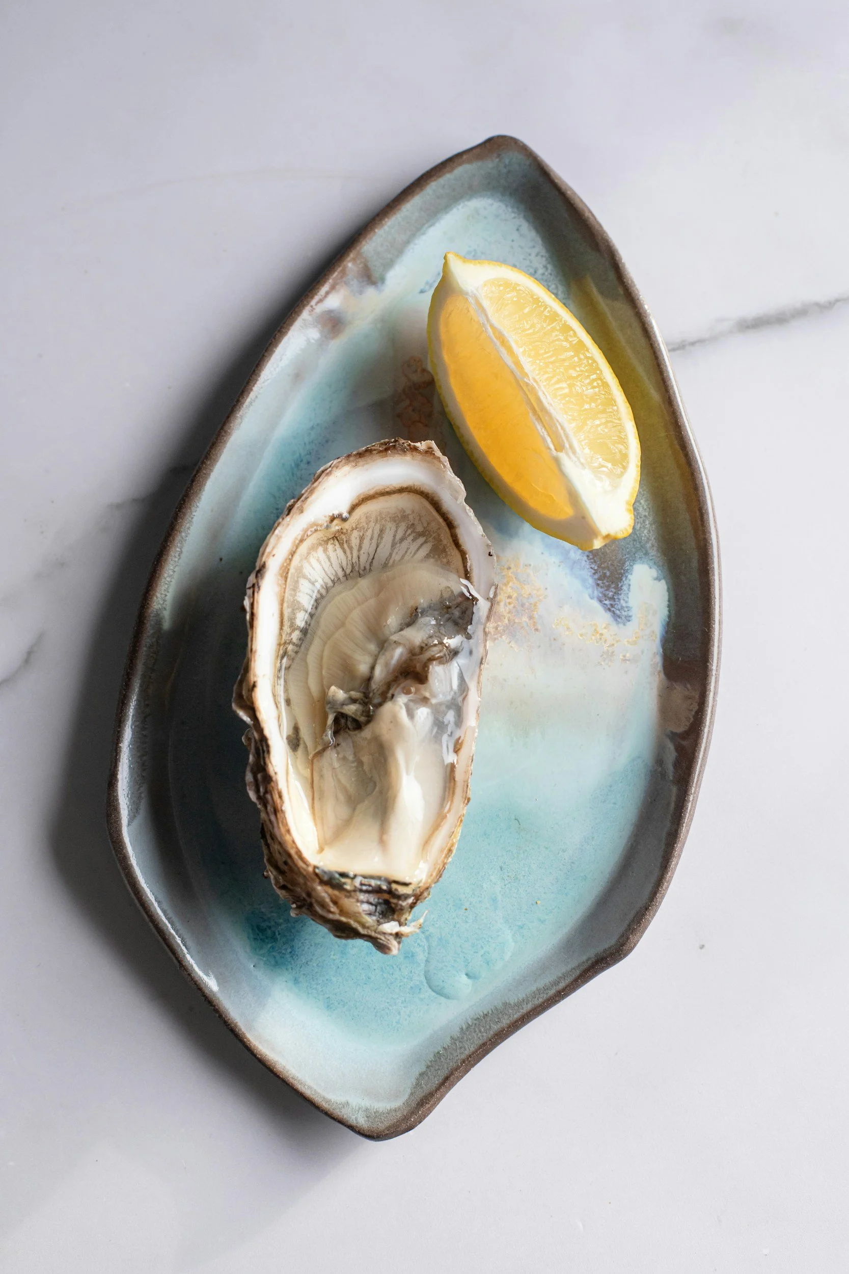 A single delicate oyster on a teal ceramic plate with a lemon wedge, showing the small, sweet Pacific-style variety