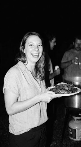 Woman smiling and holding a plate of food at a gathering with other people in the background.