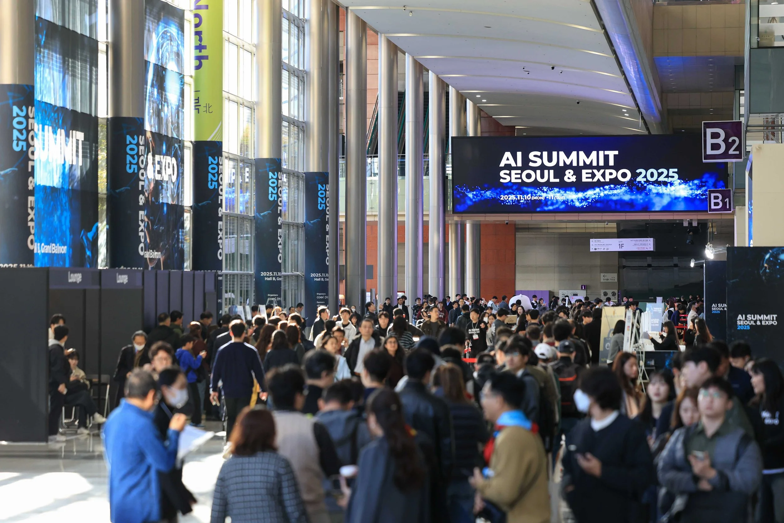 Large crowd of people attending the AI Summit Seoul & Expo 2025 inside a modern convention center with tall glass windows and digital banners.