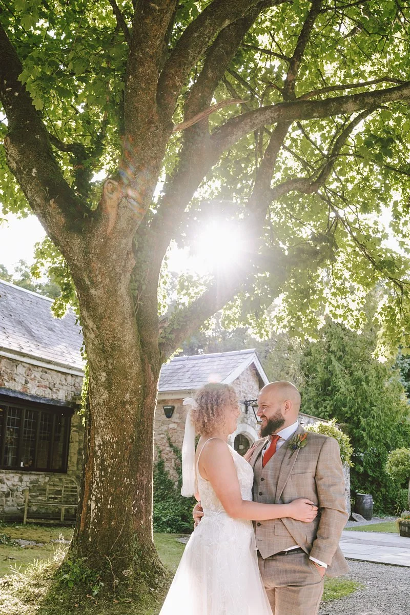Bride and groom standing in a lush green forest, holding hands. The bride is wearing a white wedding dress and veil, holding a bouquet of flowers. The groom is in a blue suit and tie. Surrounded by ferns and trees.