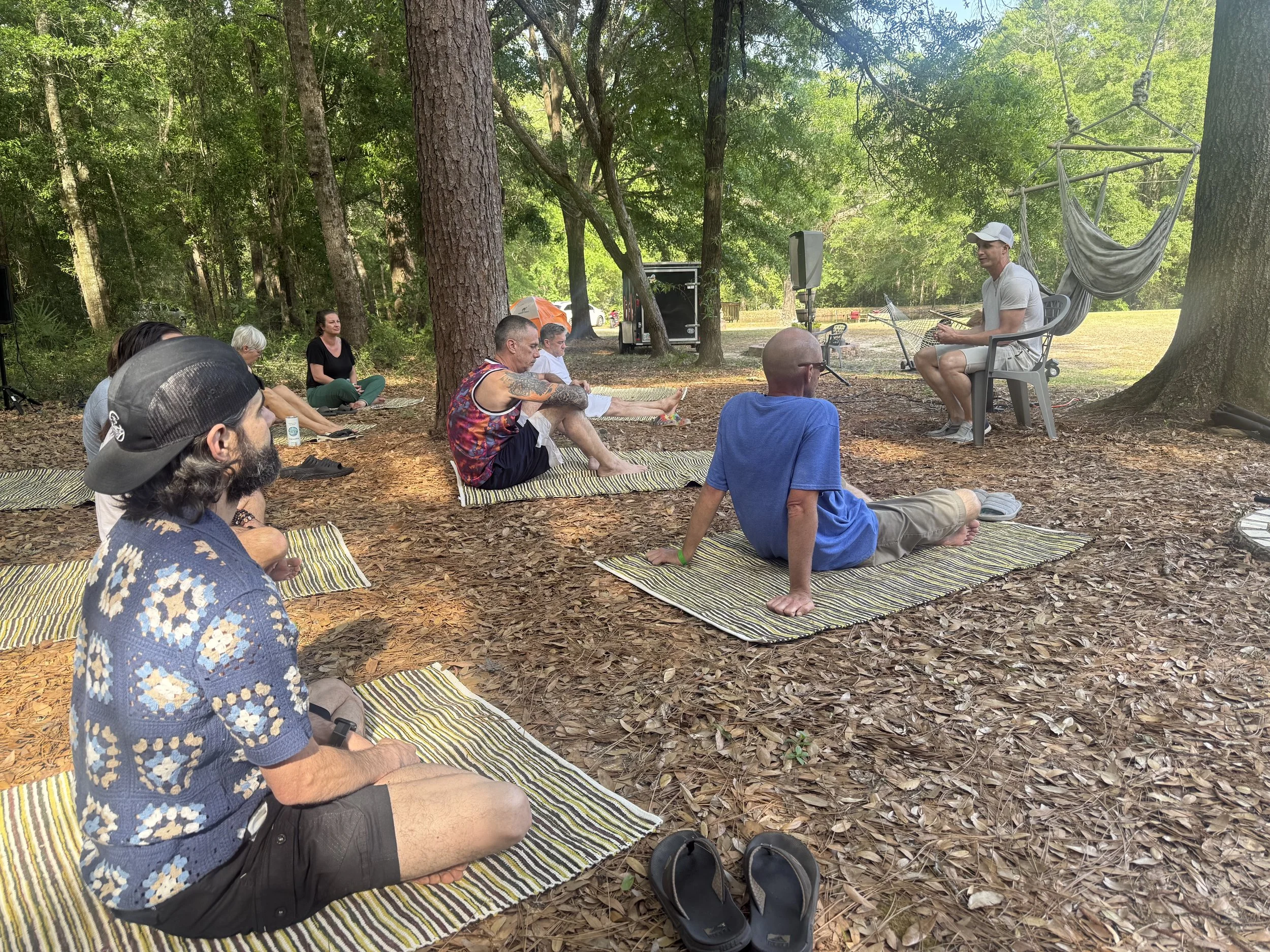 A group of people sitting on mats in a forest, attending an outdoor seminar or workshop. A man in white is seated on a chair with a hammock behind him, speaking or presenting to the group.