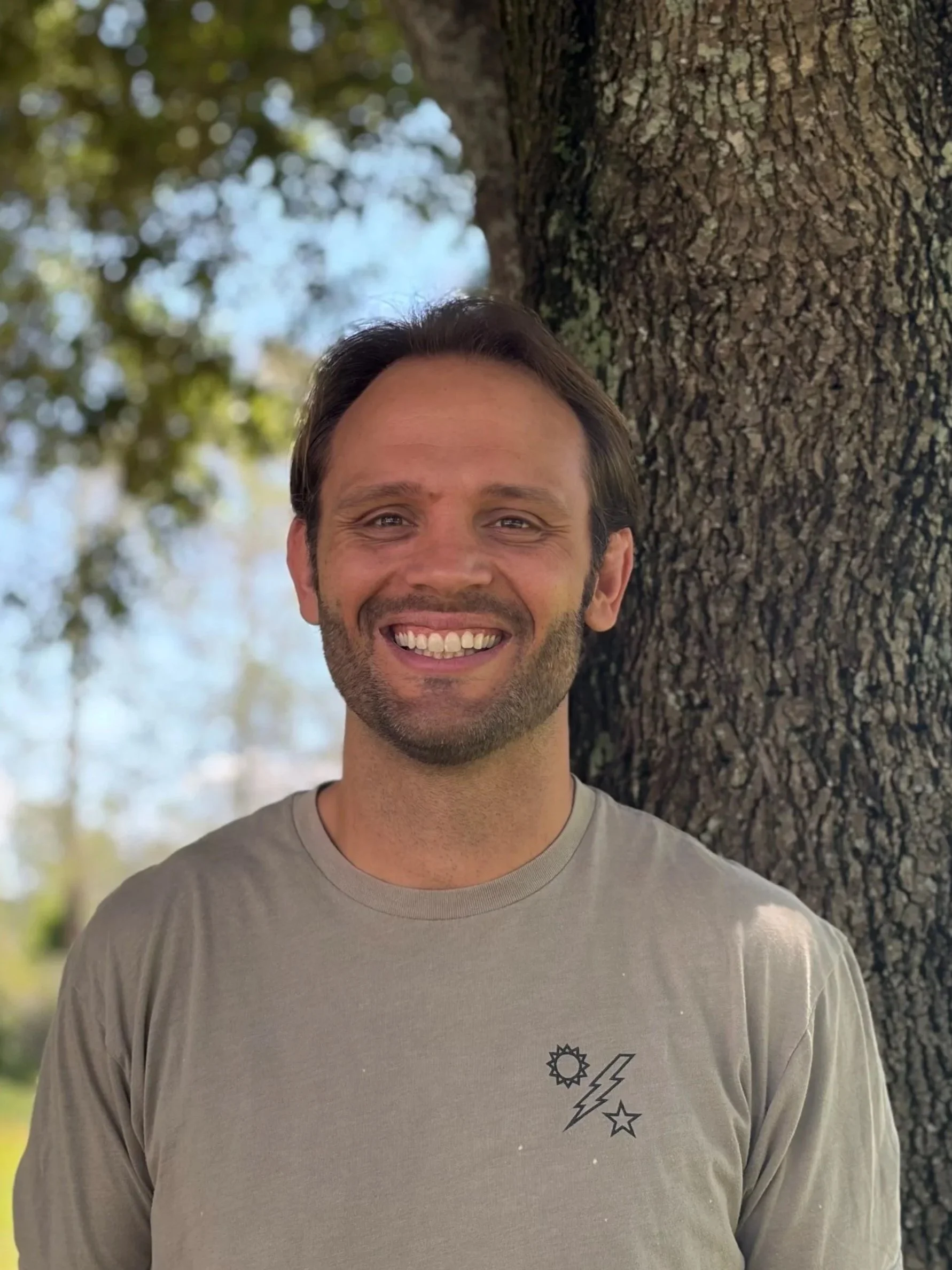 Smiling man with shoulder-length brown hair and beard, wearing a navy blue T-shirt with a large 