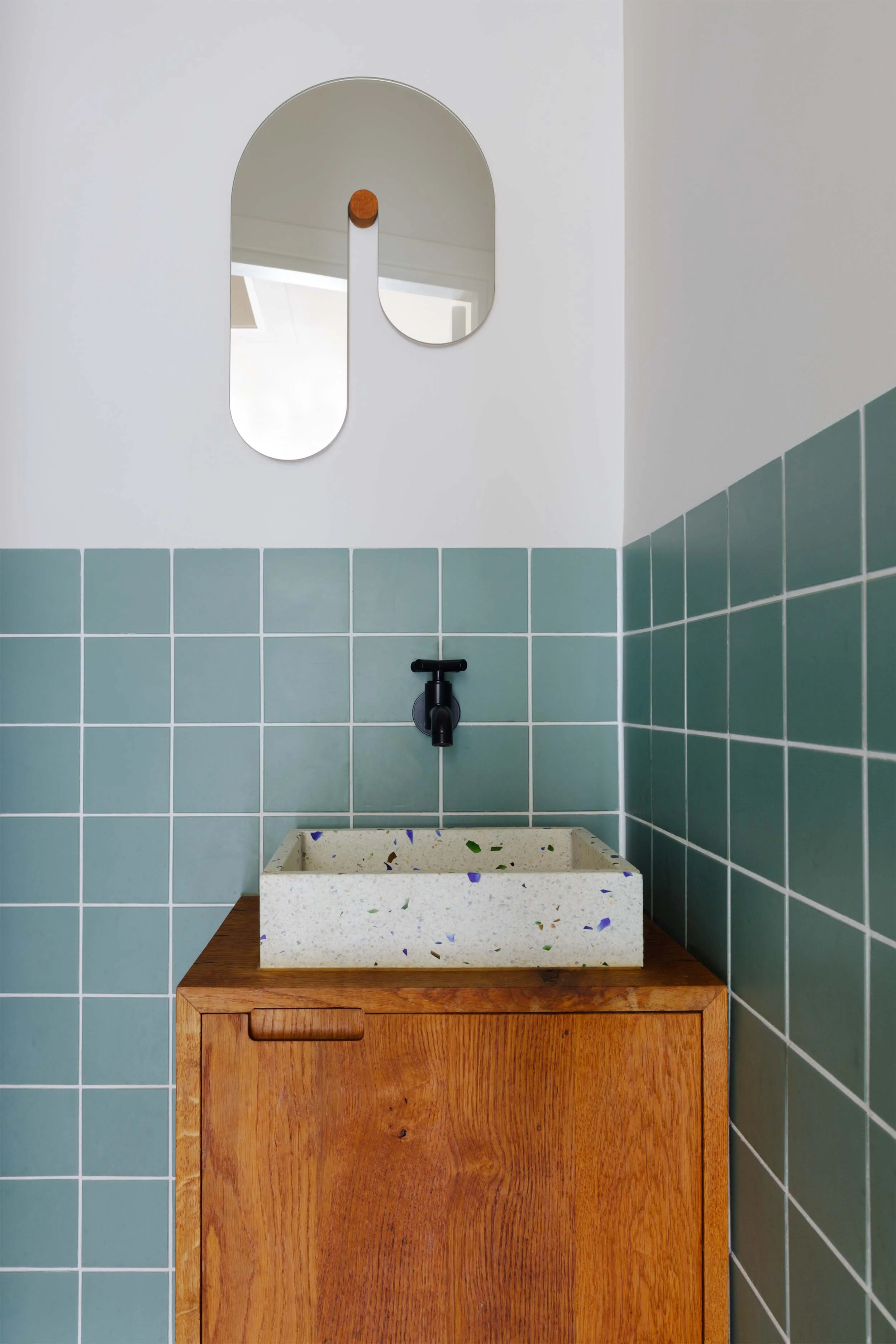 Eikenhouten toiletmeubel met zwarte kraan, terrazzo wasbak in toilet met groene tegels en ronde gebogen spiegel