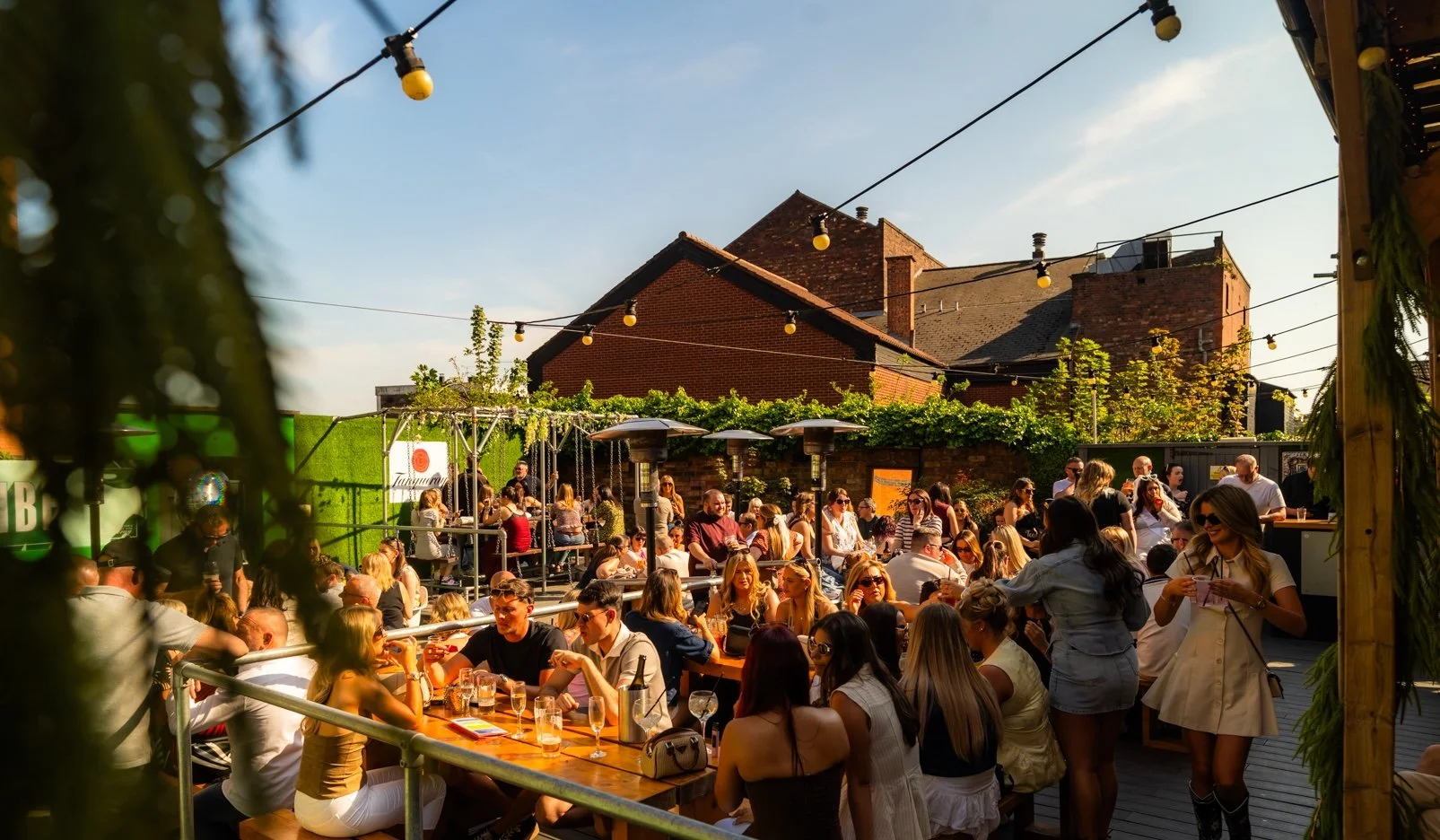 A lively outdoor gathering with many people seated at tables under string lights on a sunny day, with brick buildings and green plants in the background.