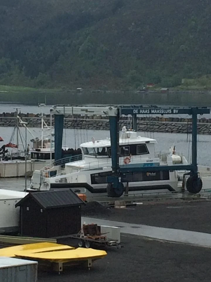 Ferge med teksten 'De Haas Maassluys BV' på. Flere båter og en liten svart hytte er synlige, med land og fjell i bakgrunnen.