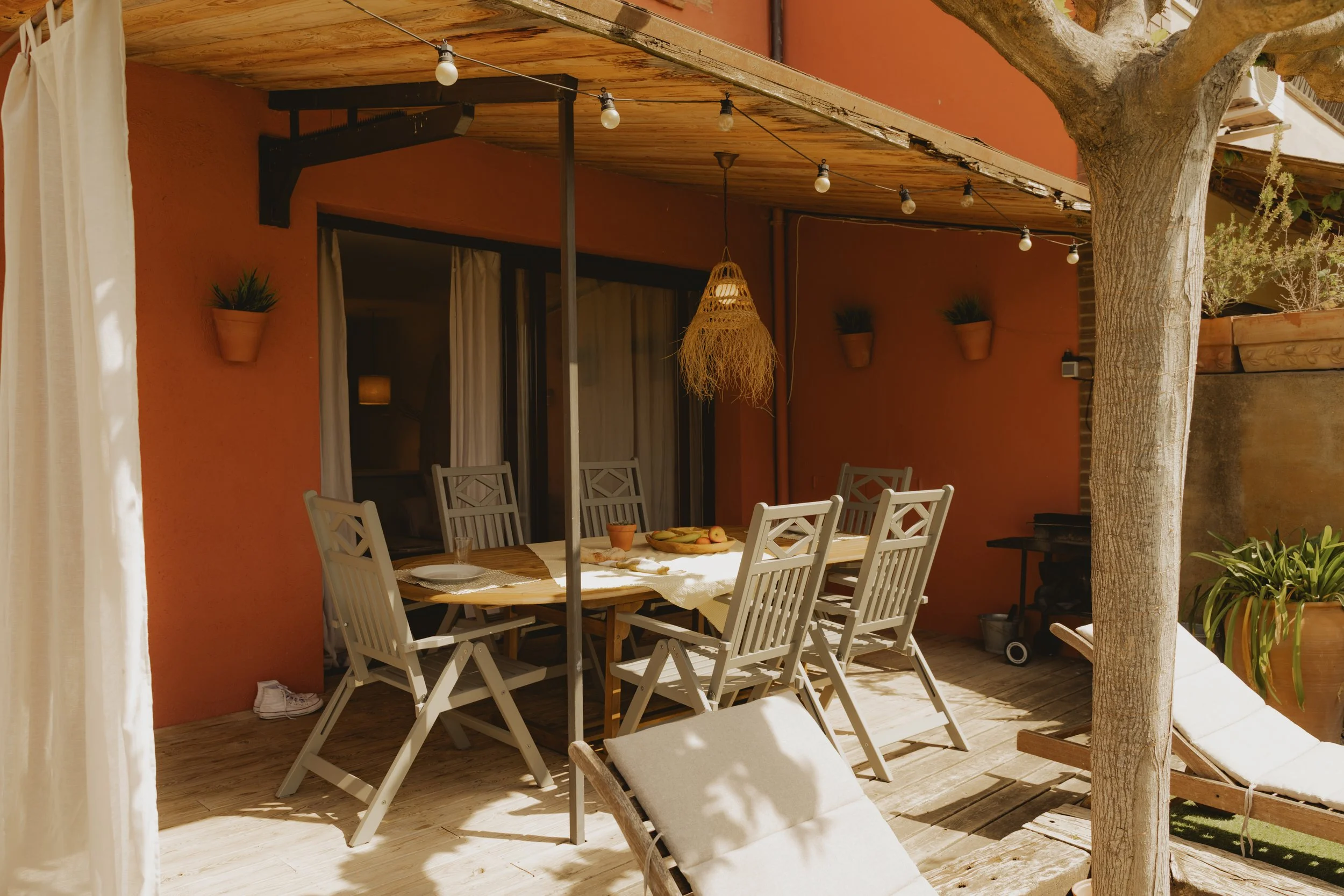 Una terraza exterior con una mesa y sillas, decorada con luces y plantas. En la mesa hay comida y utensilios, y hay una tumbona y una sombrilla en el área.