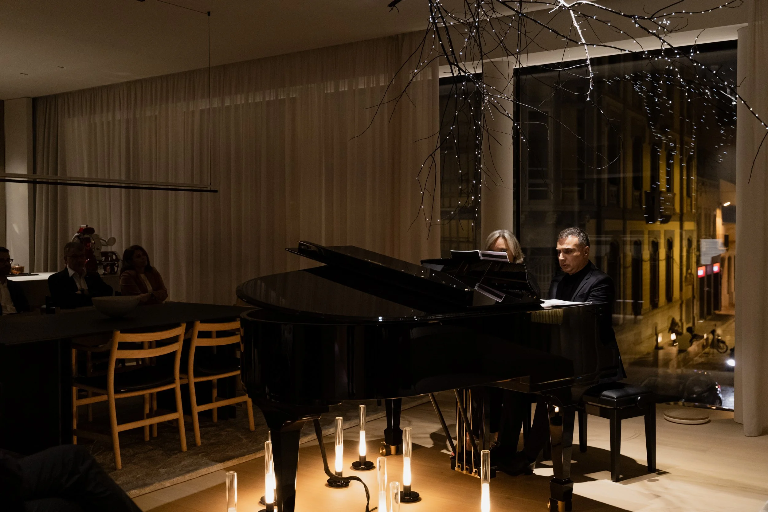 Un hombre tocando el piano en un evento nocturno con varias velas iluminando el suelo, mientras personas en el fondo observan, en un ambiente elegante y tranquilo.