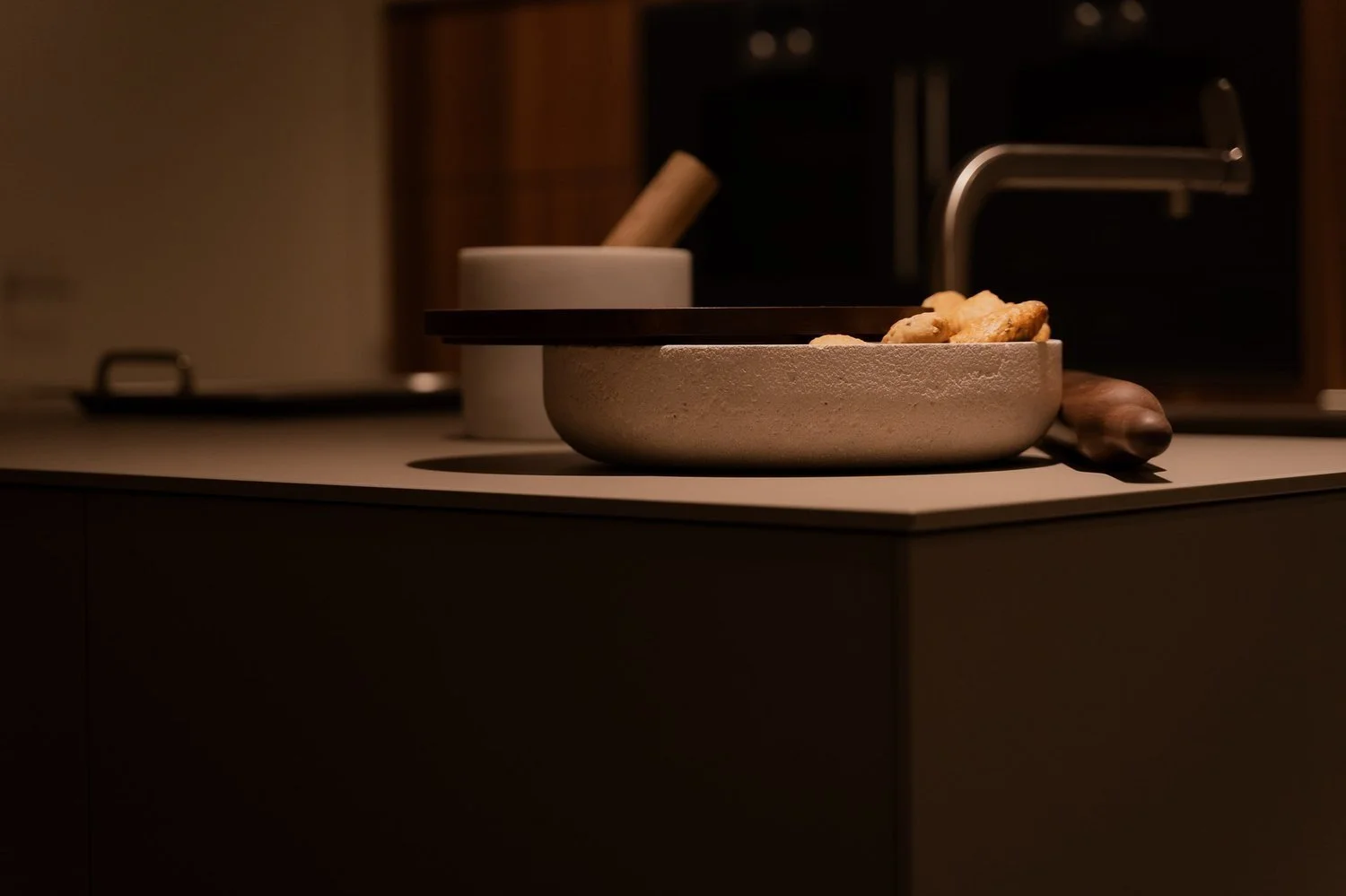 Imagen de una cocina con un sartén en la mesa, con pedazos de pan y un rodillo de amasar a su lado. En el fondo, un grifo y una taza blanca con un rodillo de madera.