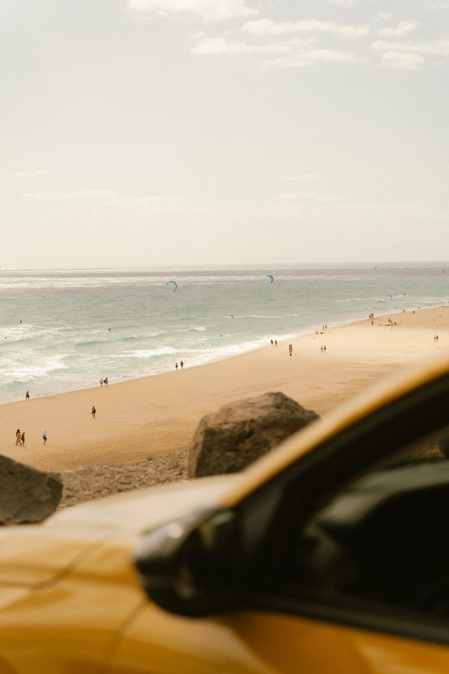 Vista de una playa con arena dorada, algunas rocas en primer plano y varias personas caminando o en el agua, con cometas en el cielo y una nube en la parte superior.