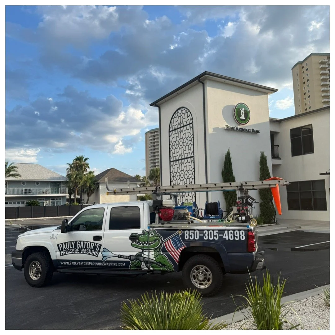 A person pressure washing the exterior wall of a Fairfield Inn & Suites hotel on a sunny day. The hotel has tan and gray panels, and a man in a blue hoodie, gray pants, and a white cap is holding a pressure washer wand directed upward. The hotel sign is visible at the top left, and there is a logo for Pauly Gator's Pressure Washing with a cartoon alligator holding a flag and a pressure wand at the bottom right corner.