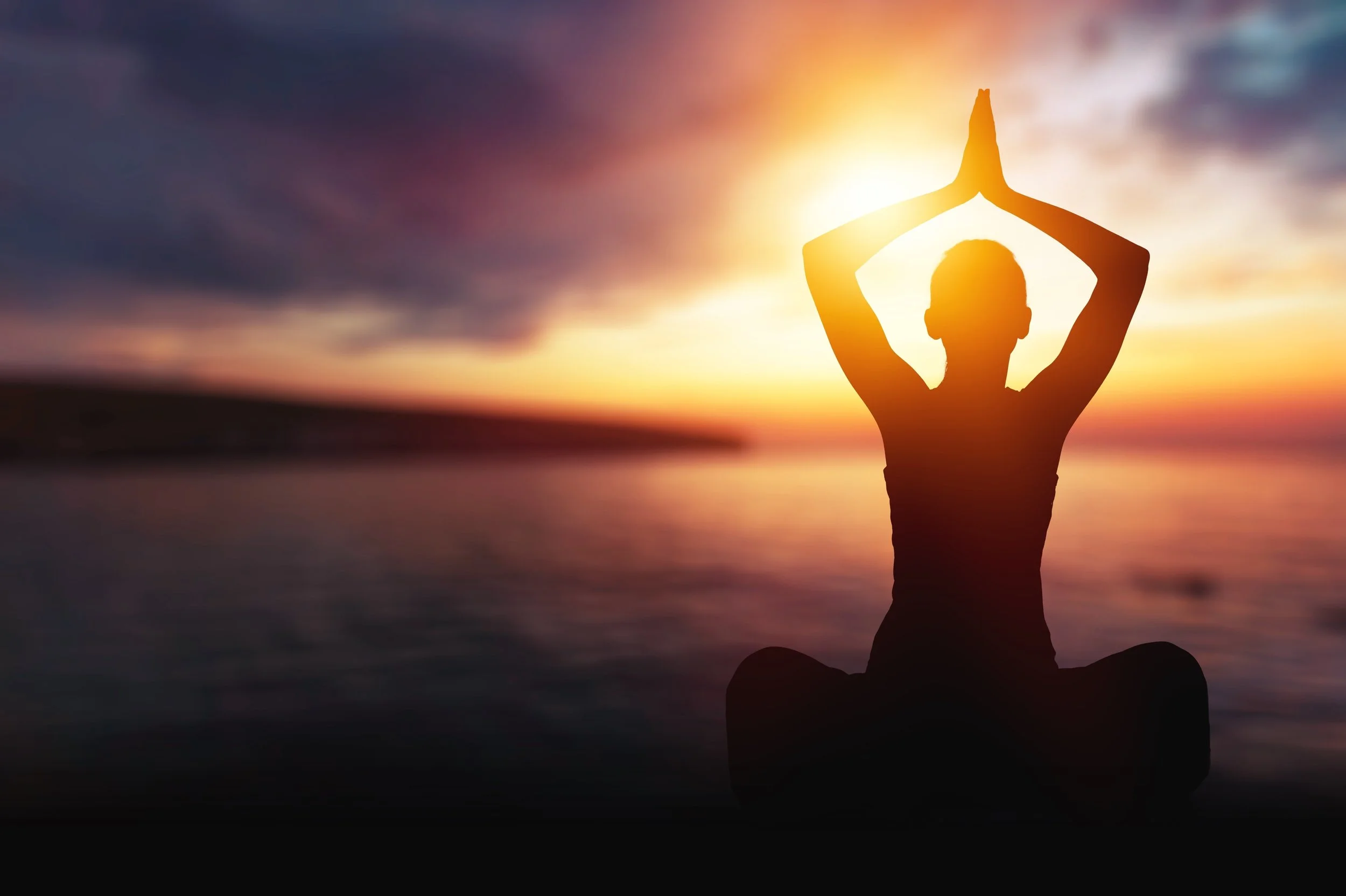 Silhouette of a person doing yoga or meditation with hands raised in a prayer pose against a sunset sky over water.