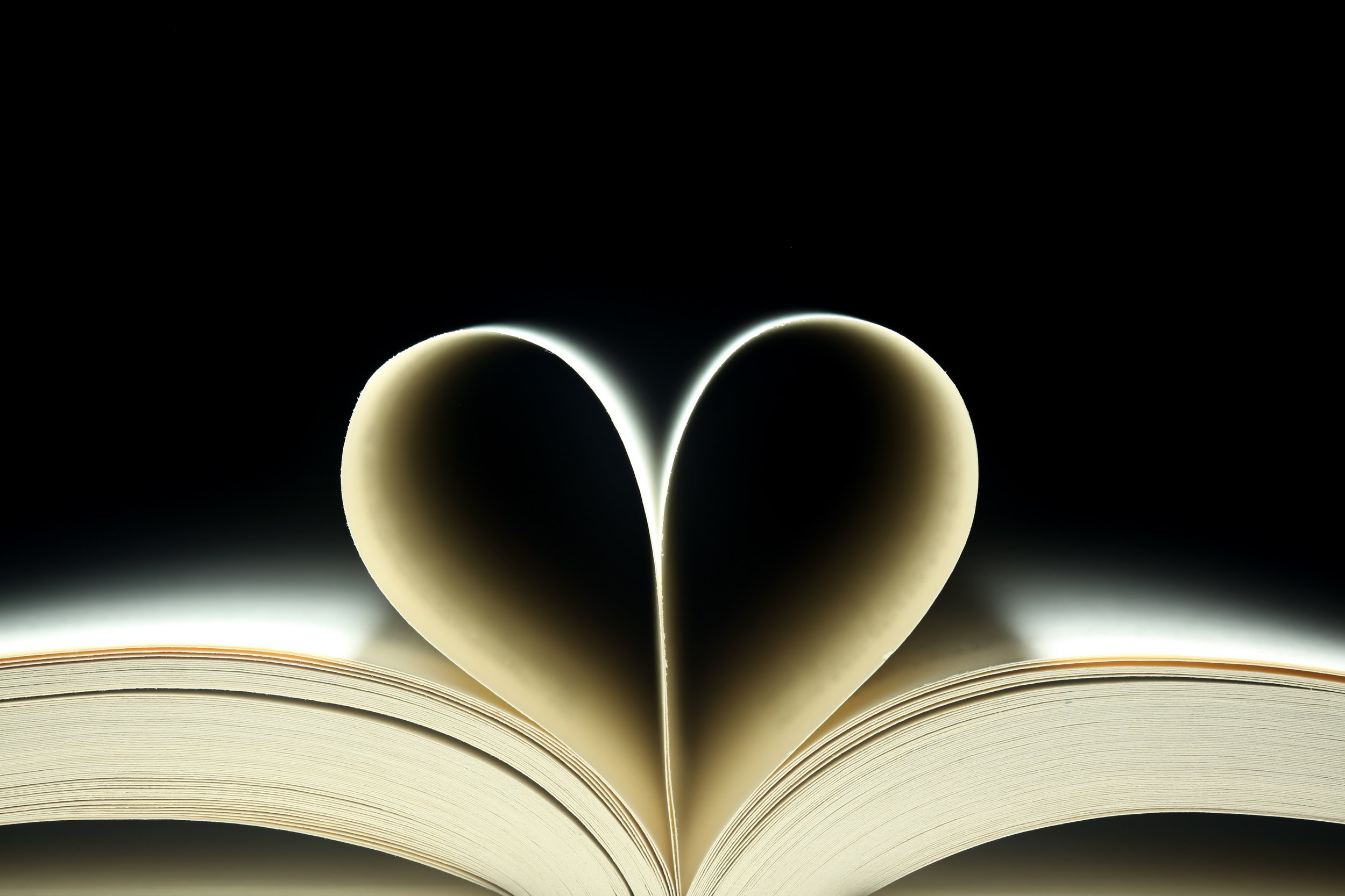 An open book with pages folded to form a heart shape, photographed against a dark background.
