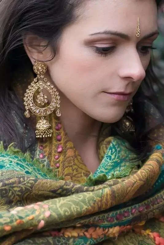 A woman wearing traditional Indian jewelry and a colorful scarf, looking downward with a serene expression.