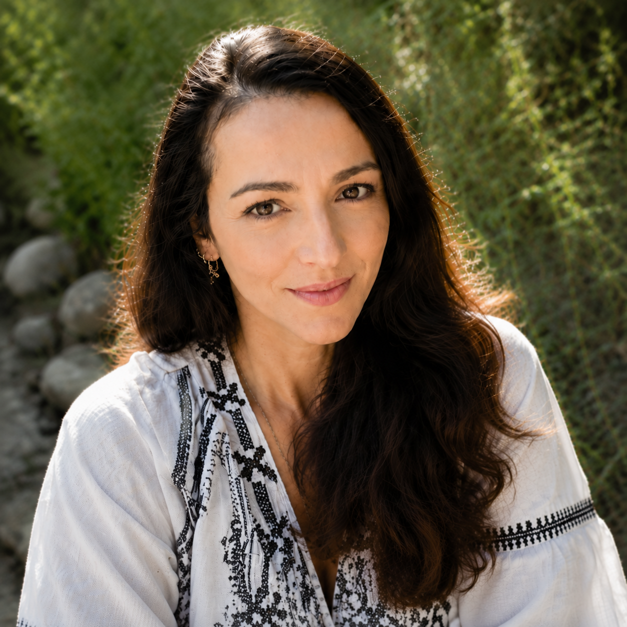 A woman with long dark hair, brown eyes, and a light complexion, smiling outdoors with greenery in the background, wearing a taupe embroidered blouse.