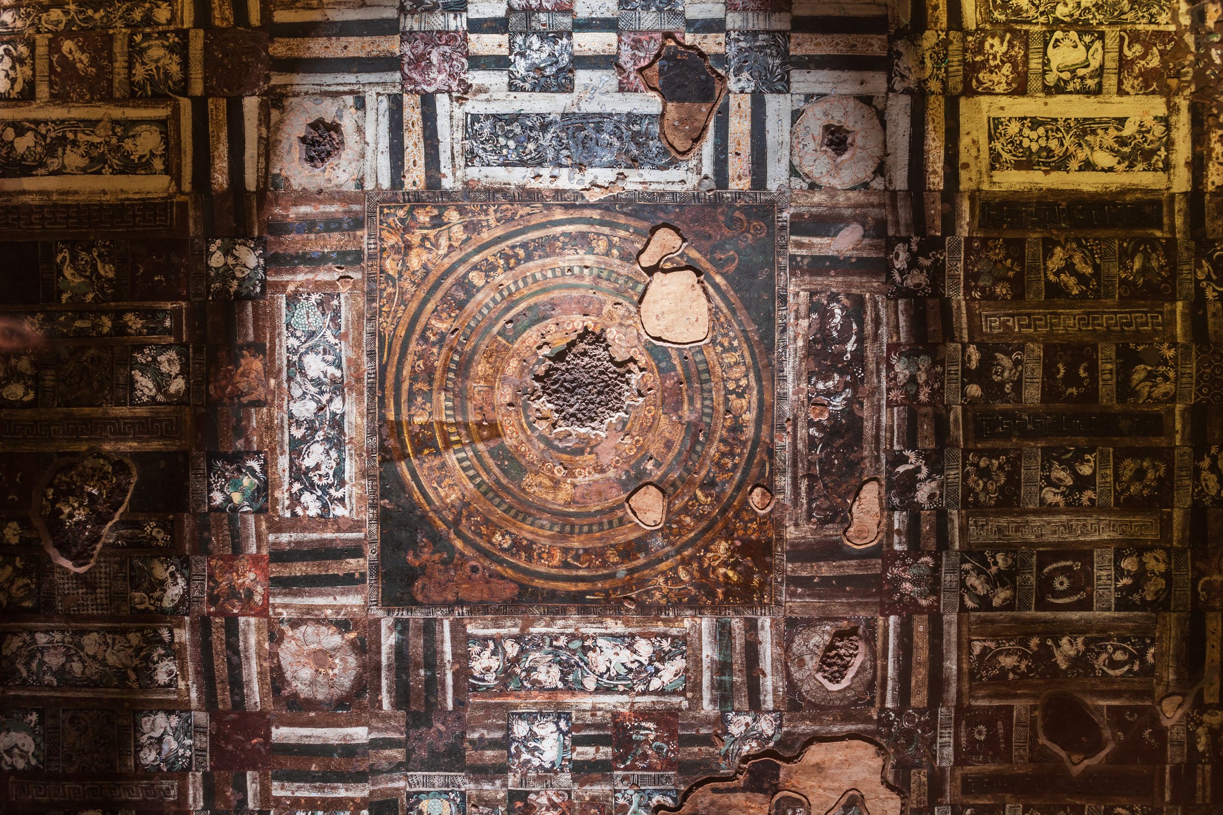 Ancient decorative mosaics in an archaeological site, with intricate geometric and floral patterns, some damaged with missing tiles.