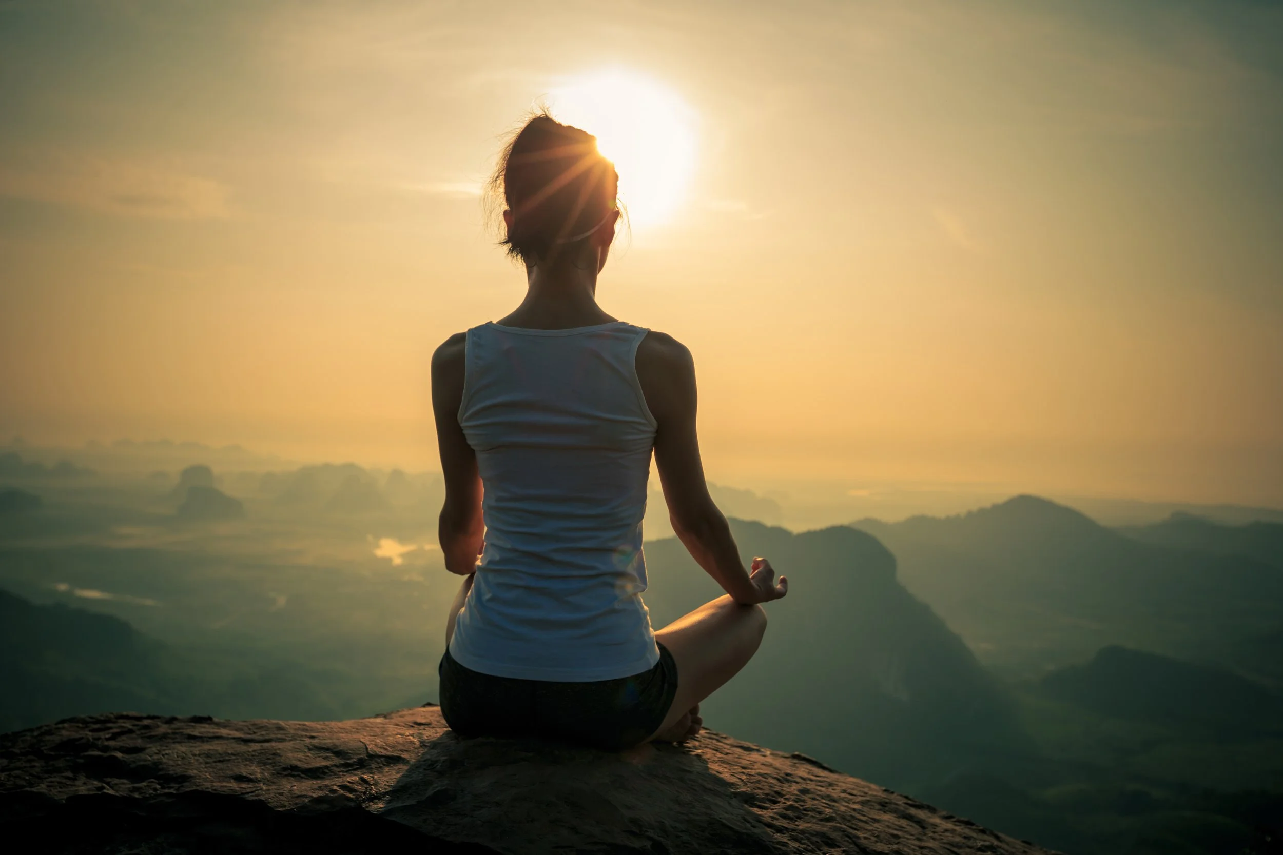 A person practicing yoga in a seated meditation pose on a rock during sunrise or sunset, overlooking a mountain landscape.