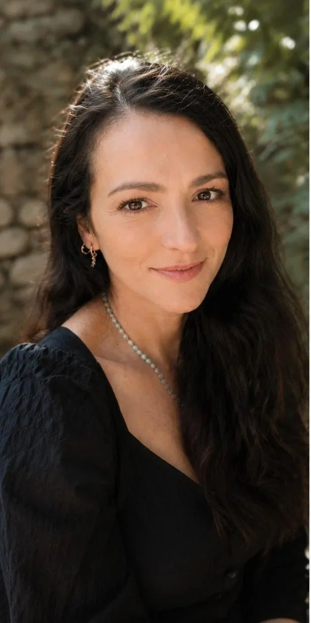 A woman with long dark hair, wearing earrings and mala beads, smiling outdoors with a stone wall and green foliage in the background.