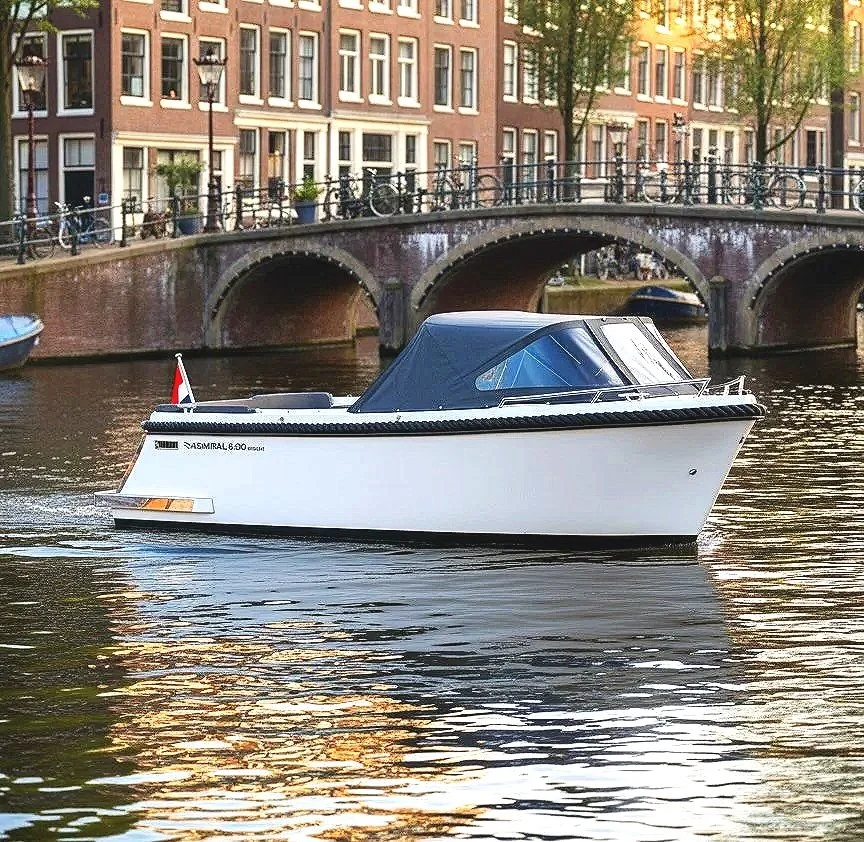Een lichtgrijze Admiral 600 sloep vaart door een Amsterdam met een brug en historische gebouwen op de achtergrond. De boot heeft een luxe uitstraling met Wormer Watersport logo.