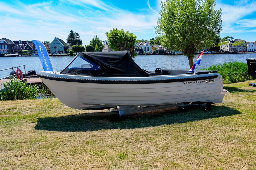 Lichtgrijze Admiral 565 sloep met zwarte kap op de kant langs het water