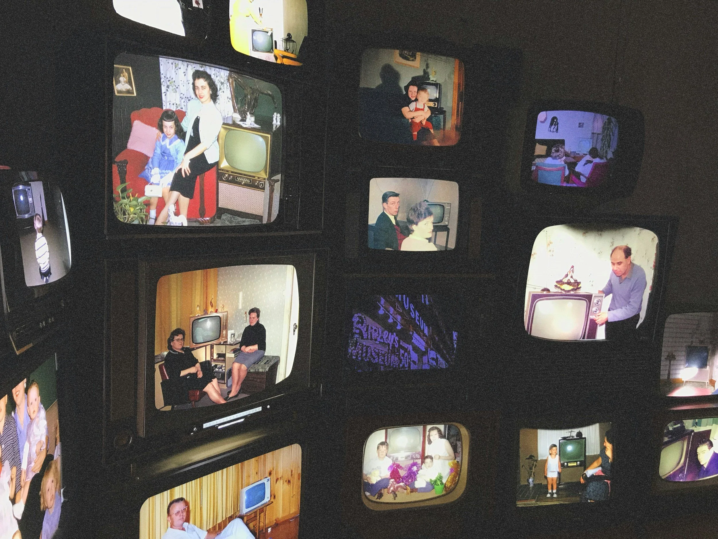 Several vintage television screens showing black and white and color images of people, including children in casual clothing, adults sitting at desks, and a woman and girl sitting on a chair, with a dark background.
