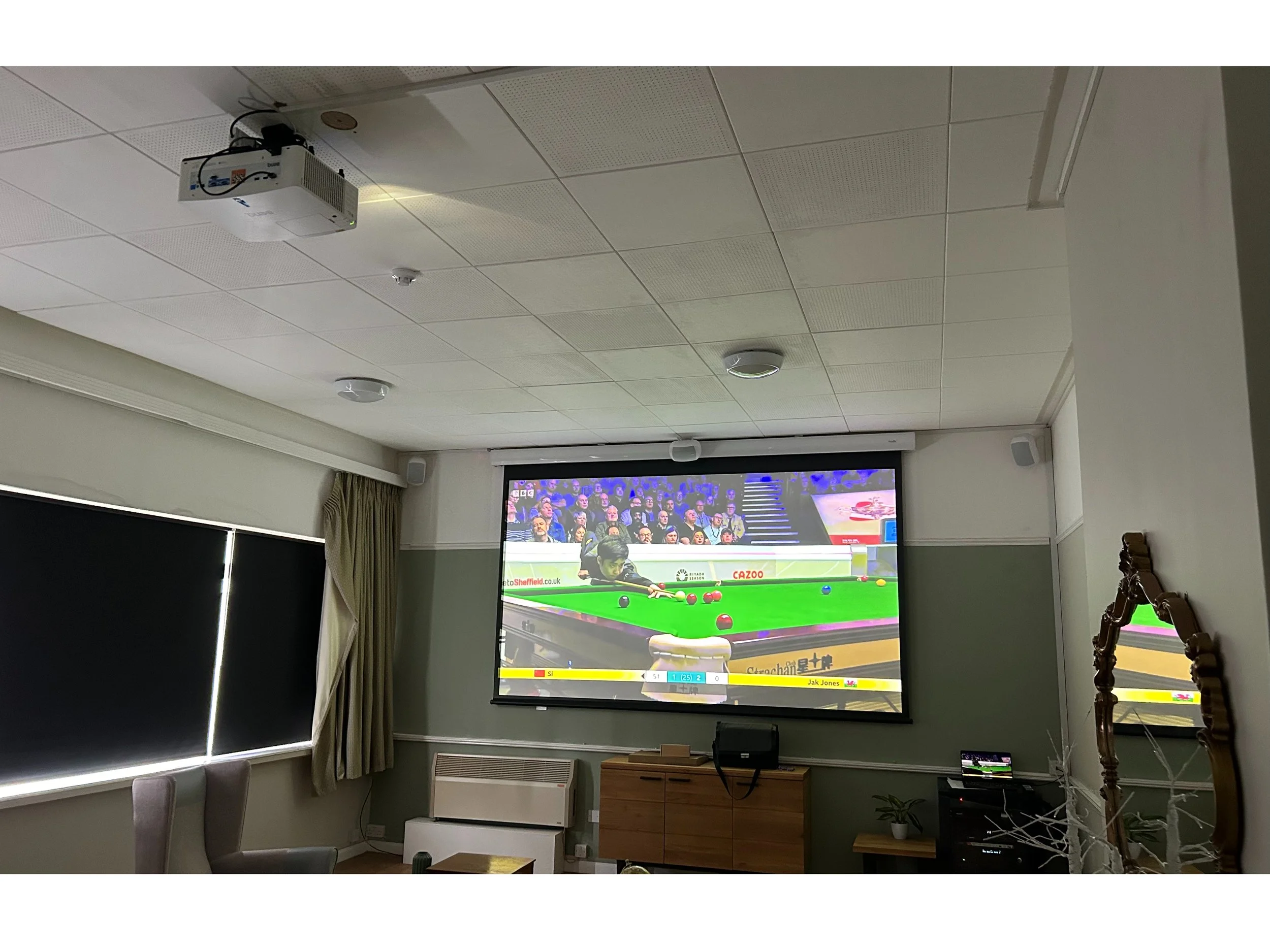 Indoor room with a ceiling-mounted projector displaying a snooker game, window with blinds, curtains, and some furniture including a plant and a wooden carved chair.