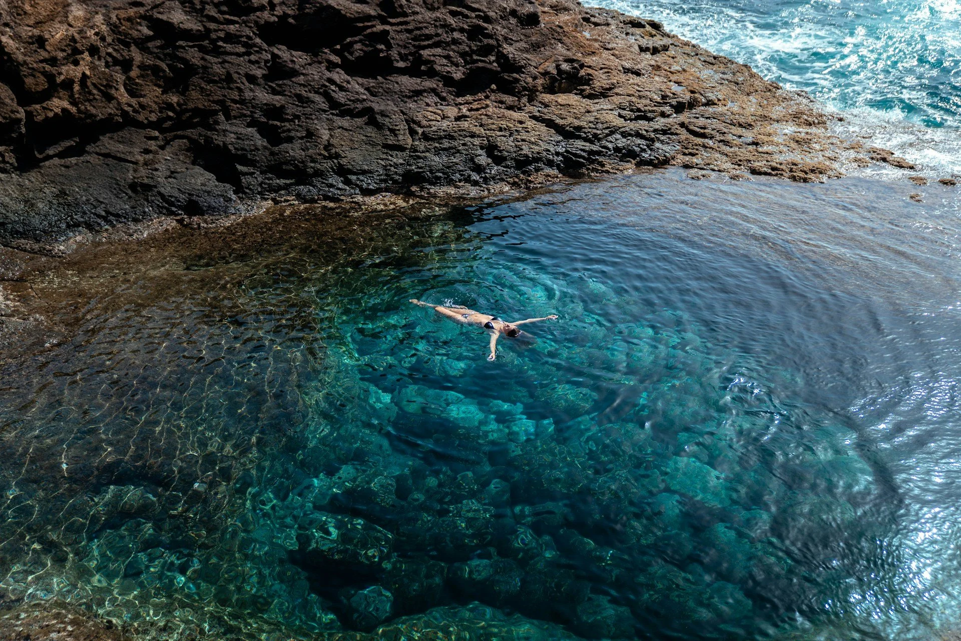 A woman floating on her back, arms outstretched in a big body of ocean by rocky edges, the sea deep turquoise and clear