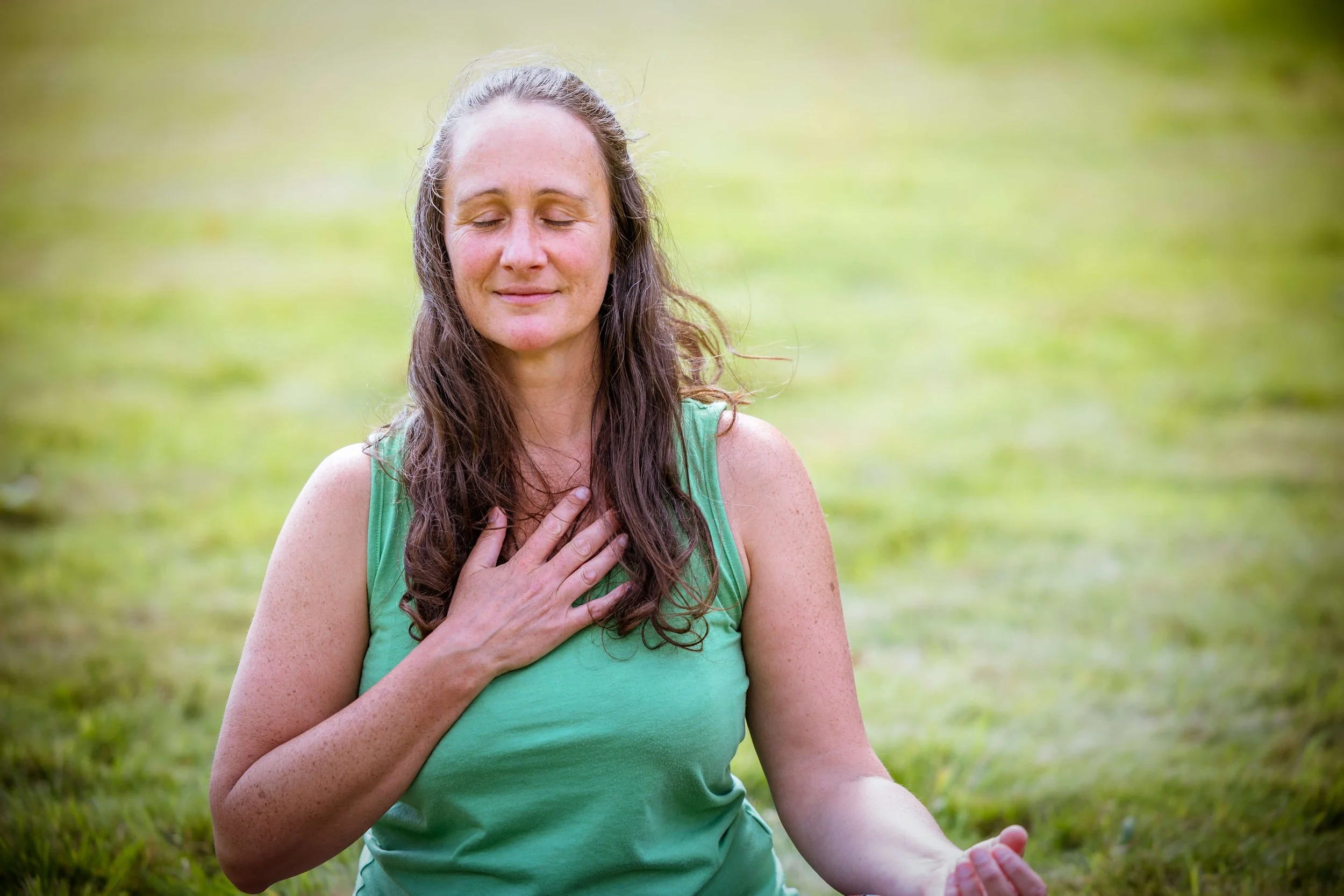 Person sitting on the grass in a green top with eyes closed and head over the heart in meditation