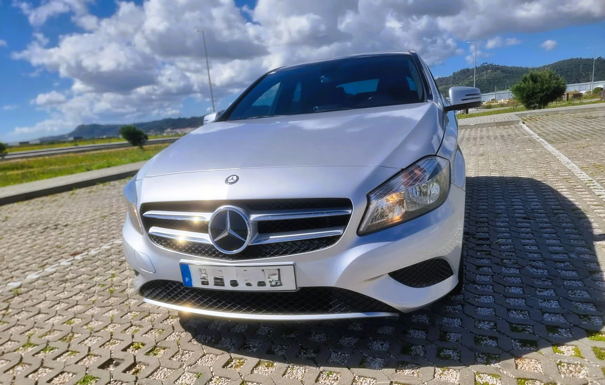 Carro prata da Mercedes-Benz estacionado em um estacionamento ao ar livre, com céu parcialmente nublado e montanhas ao fundo.