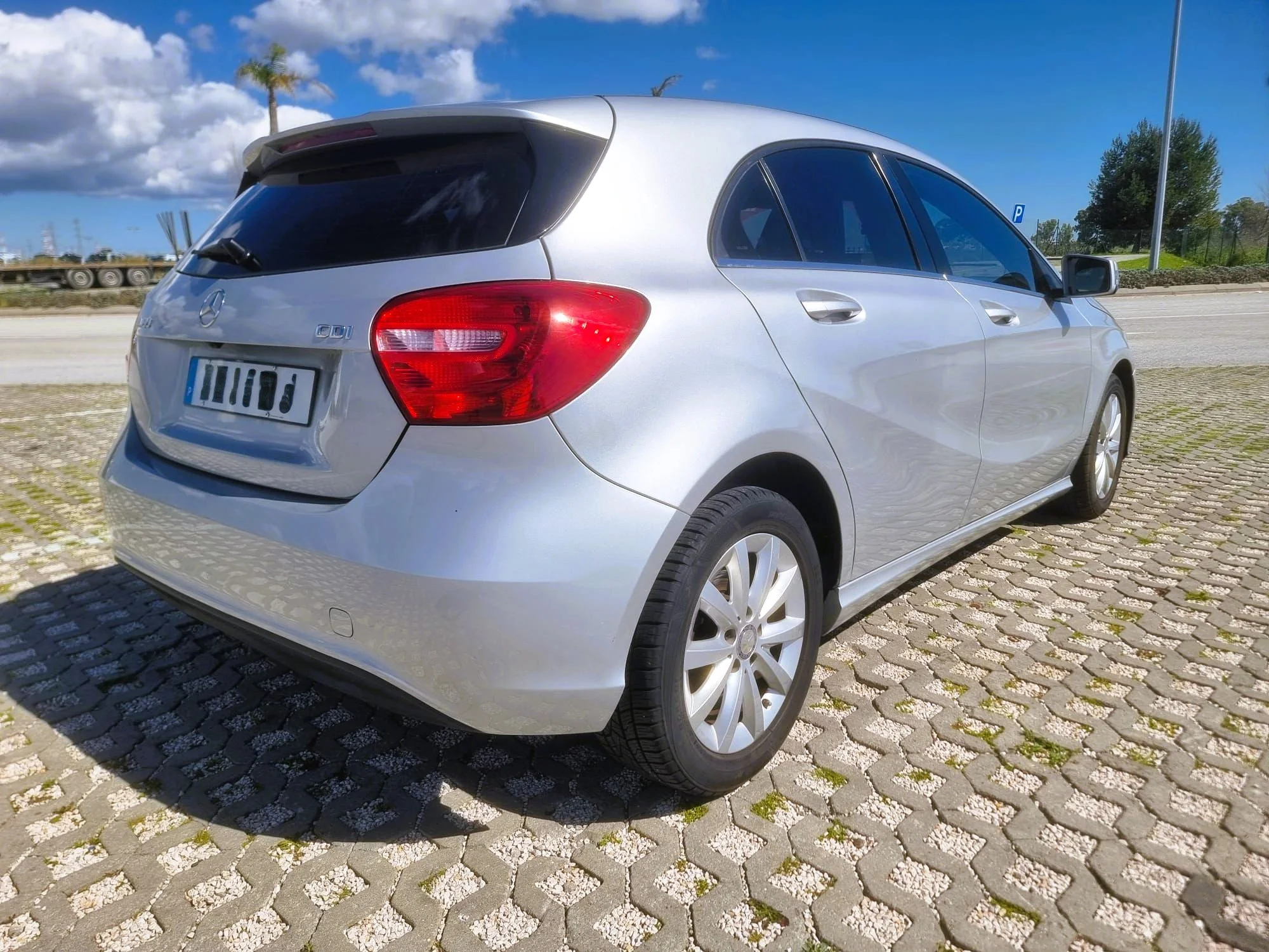 Carro prata estacionado em calçada de pedras, ao lado de uma rua, com árvores, céu azul e nuvens ao fundo.