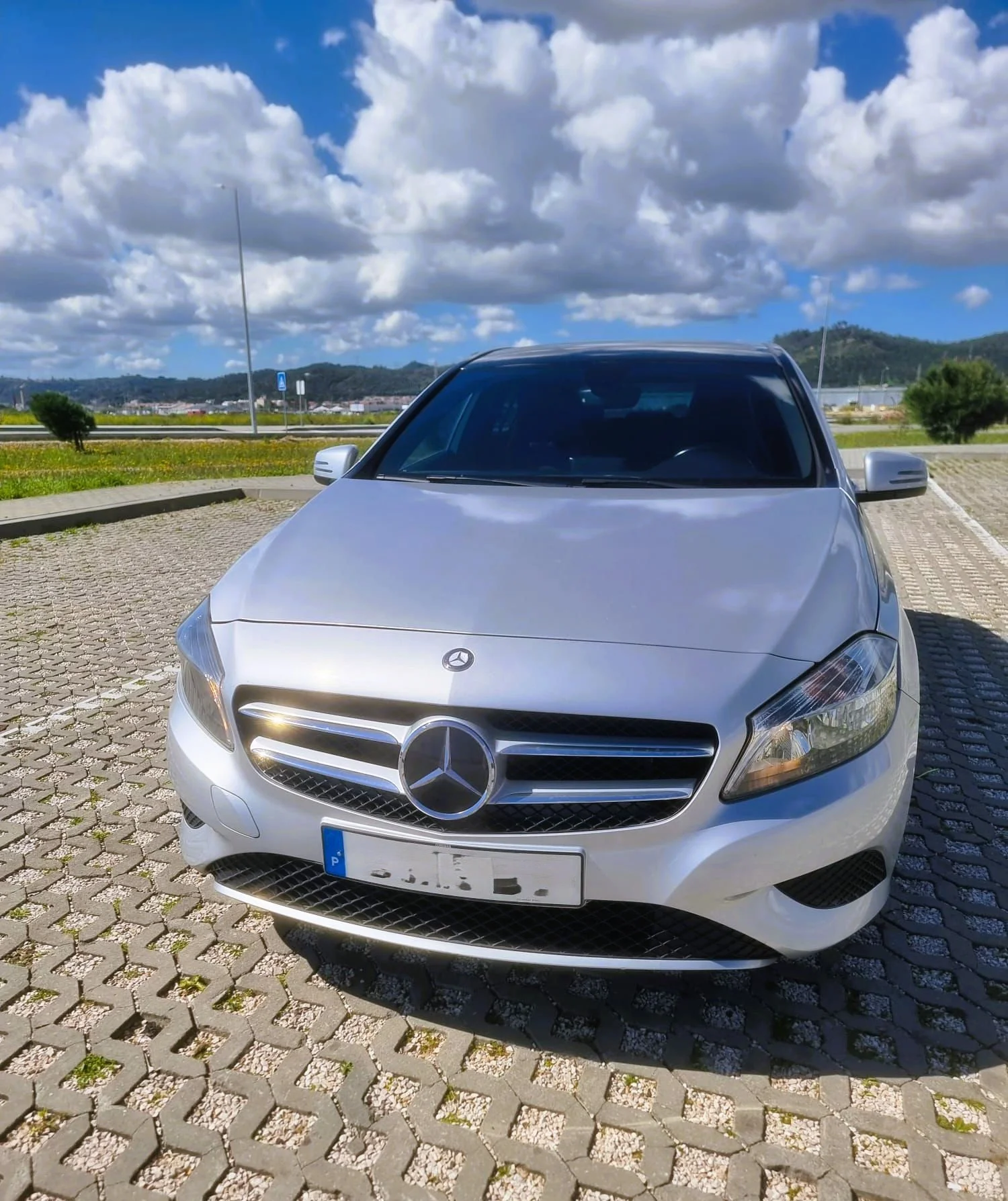 Carro prata Mercedes-Benz estacionado na entrada de um estacionamento ao ar livre, com céu parcialmente nublado ao fundo.