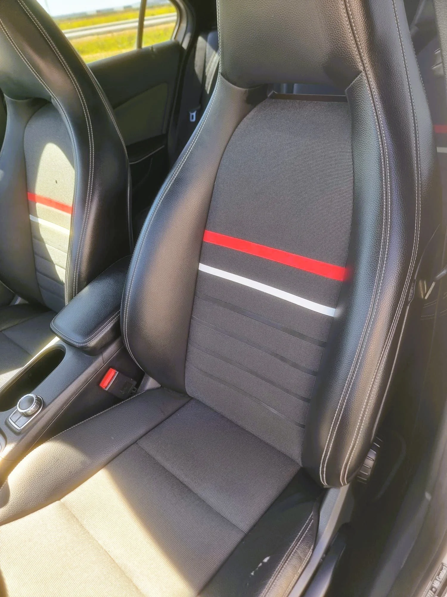 Assento de carro com tecido preto, detalhes em couro, faixa vermelha, branca e preta no encosto, com cinto de segurança ao lado.