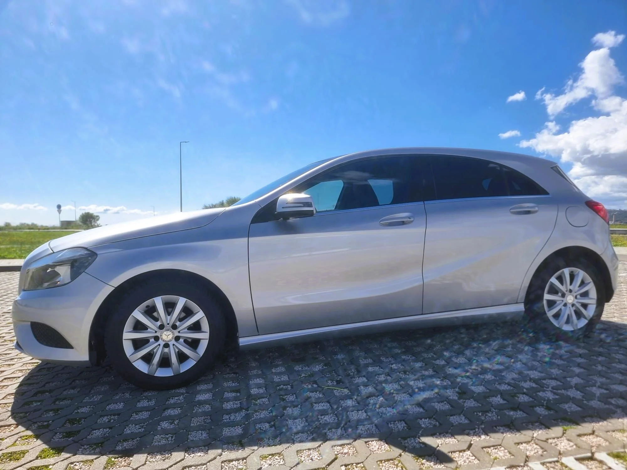 Carro sedan prata estacionado em rua de paralelepípedos sob céu azul com algumas nuvens.