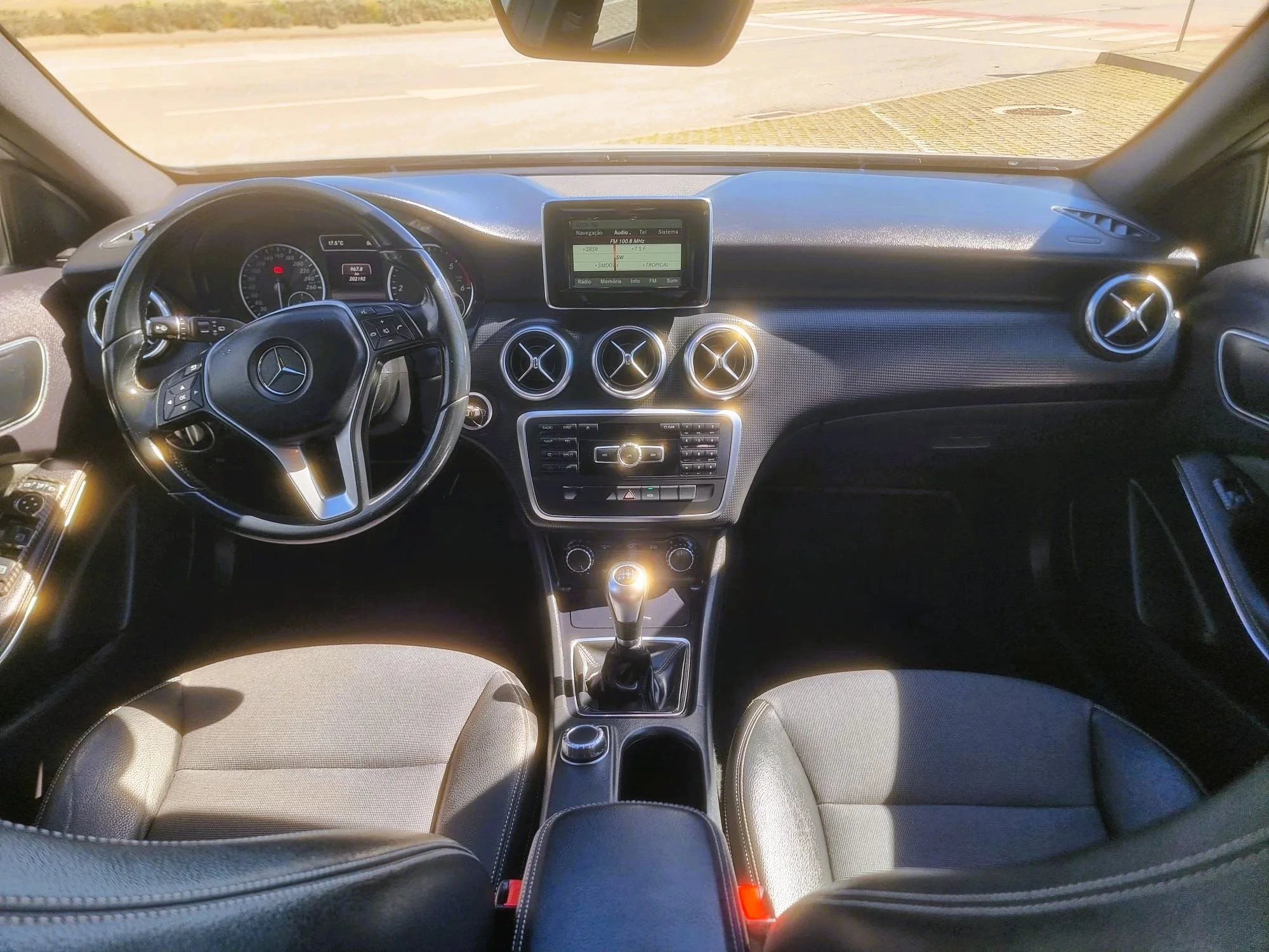 Interior de carro de luxo com assentos de couro, painel com tela de navegação, volante com comandos, centro de entretenimento com botões, controles de rádio, ar-condicionado e câmbio manual.